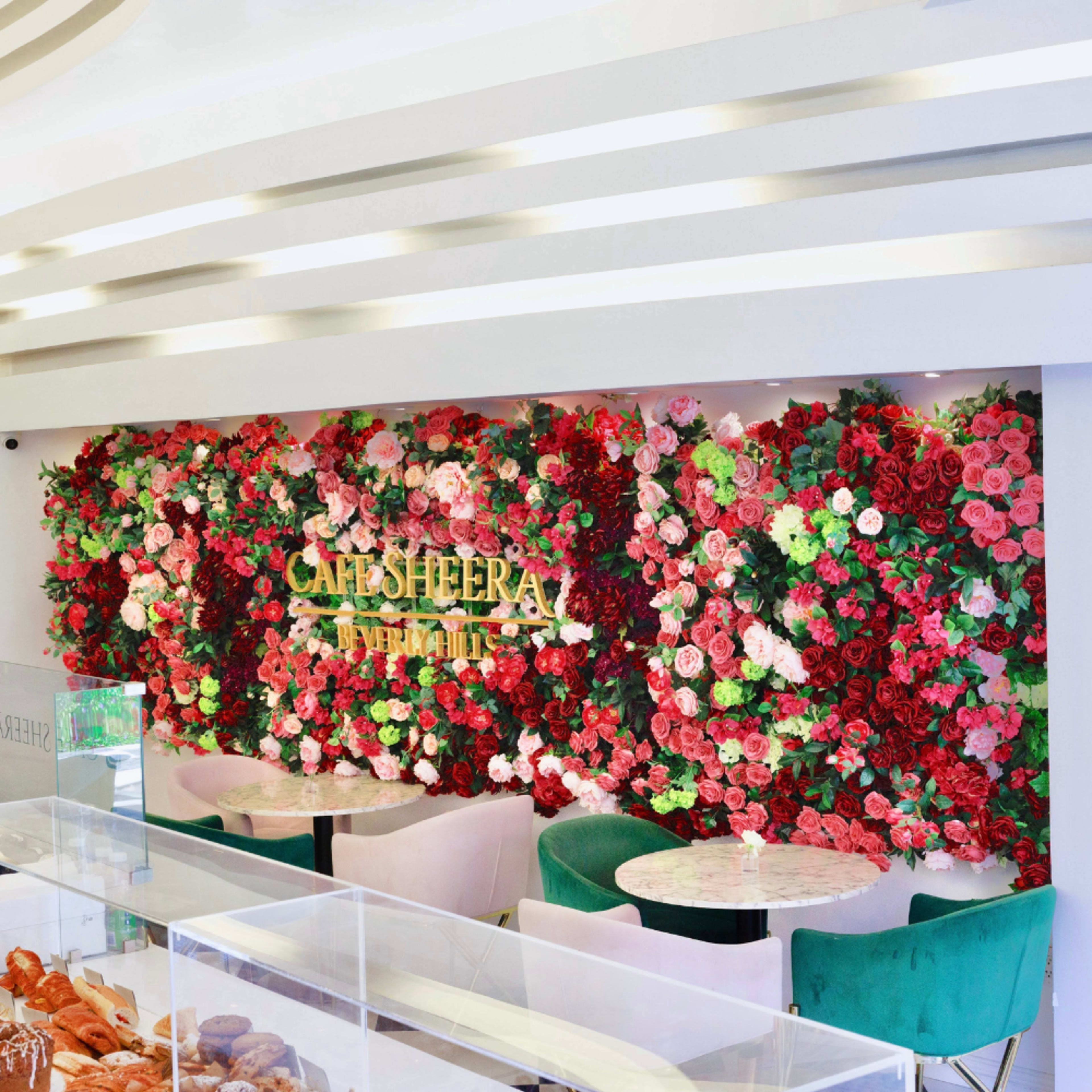 A cafe interior featuring a wall adorned with an arrangement of vibrant artificial flowers and a display case with pastries in front of green upholstered seating.