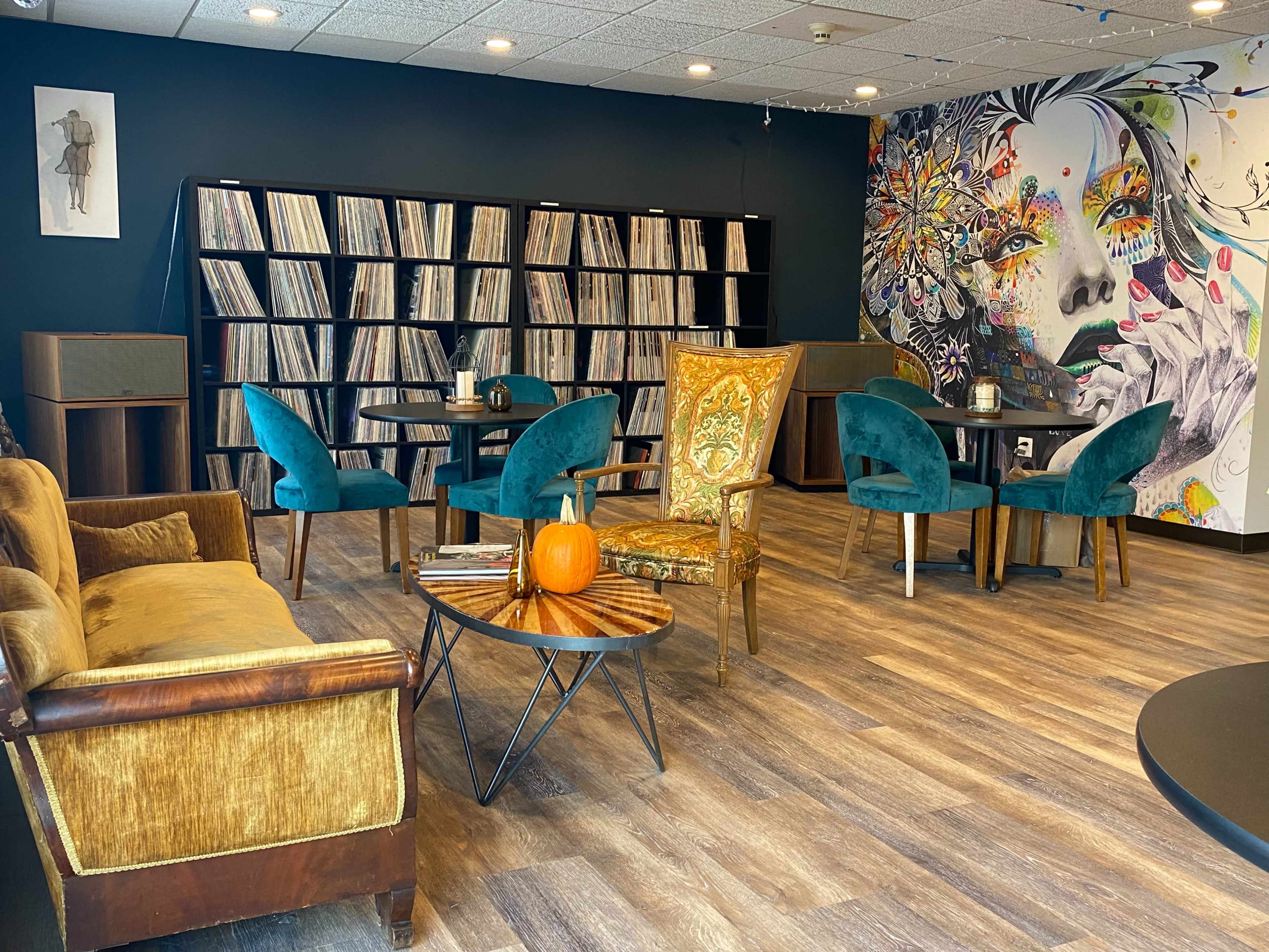 The room features a combination of vintage furniture, including a gold-accented chair and a brown couch, surrounded by shelves filled with vinyl records.