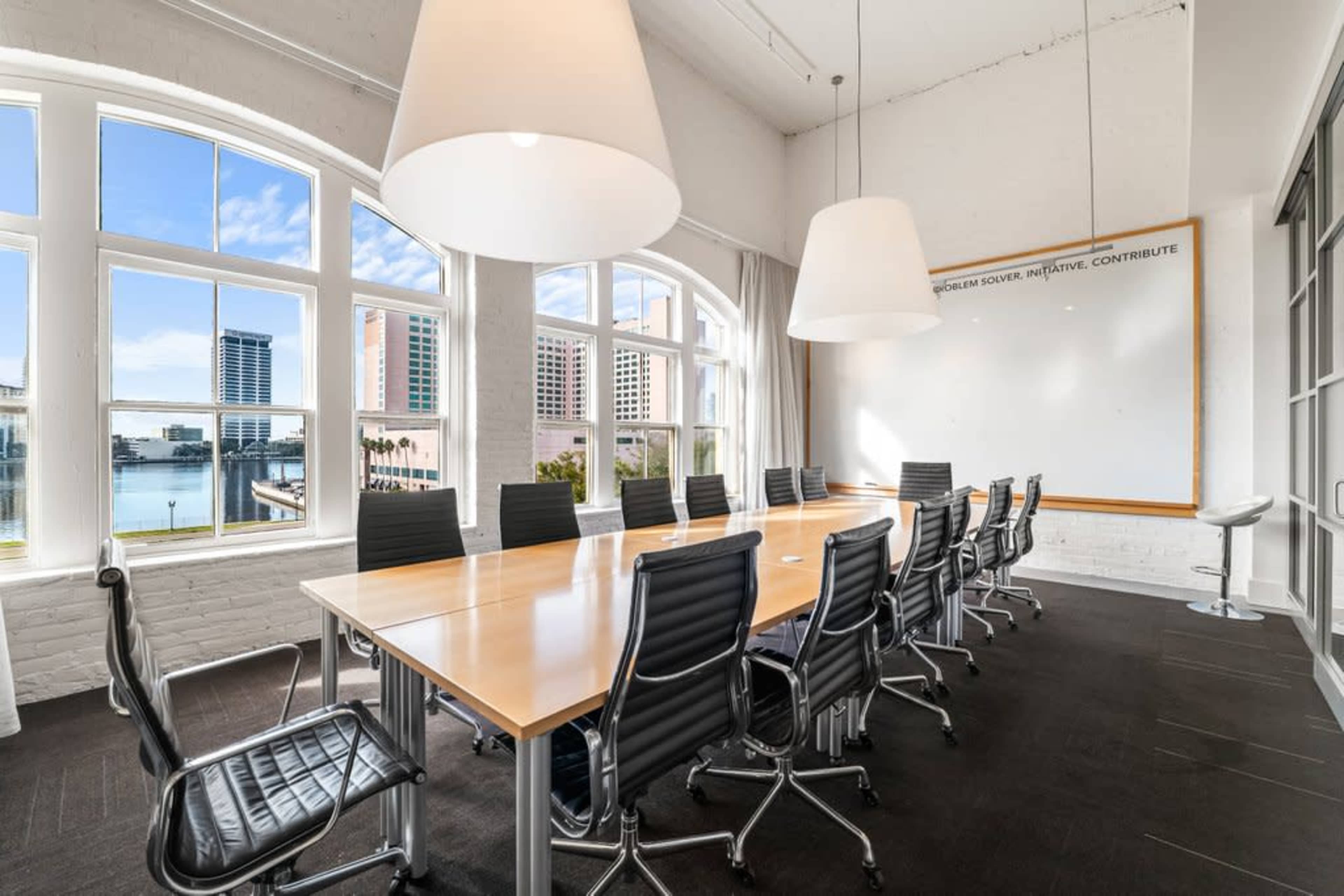A conference room featuring a long wooden table surrounded by black ergonomic chairs, with large windows providing a view of a body of water and cityscape.