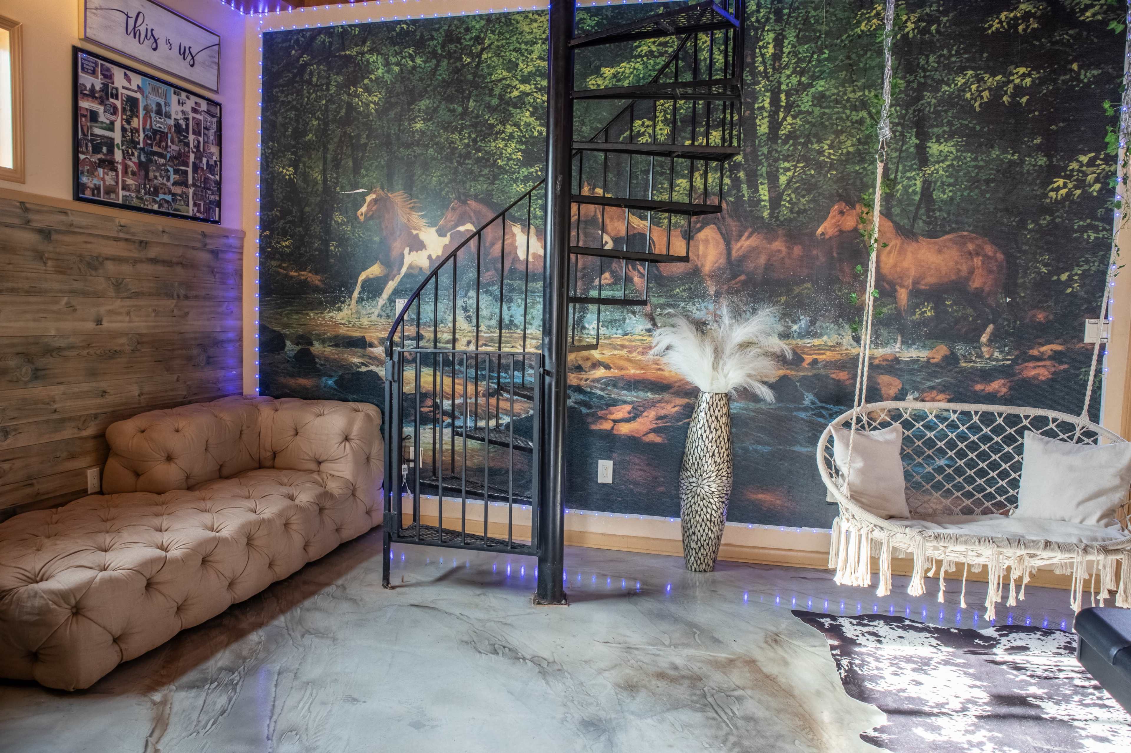 The image shows a cozy room featuring a spiral staircase, a tufted couch, and a hanging chair, with a large mural of horses in a forested setting on the wall.