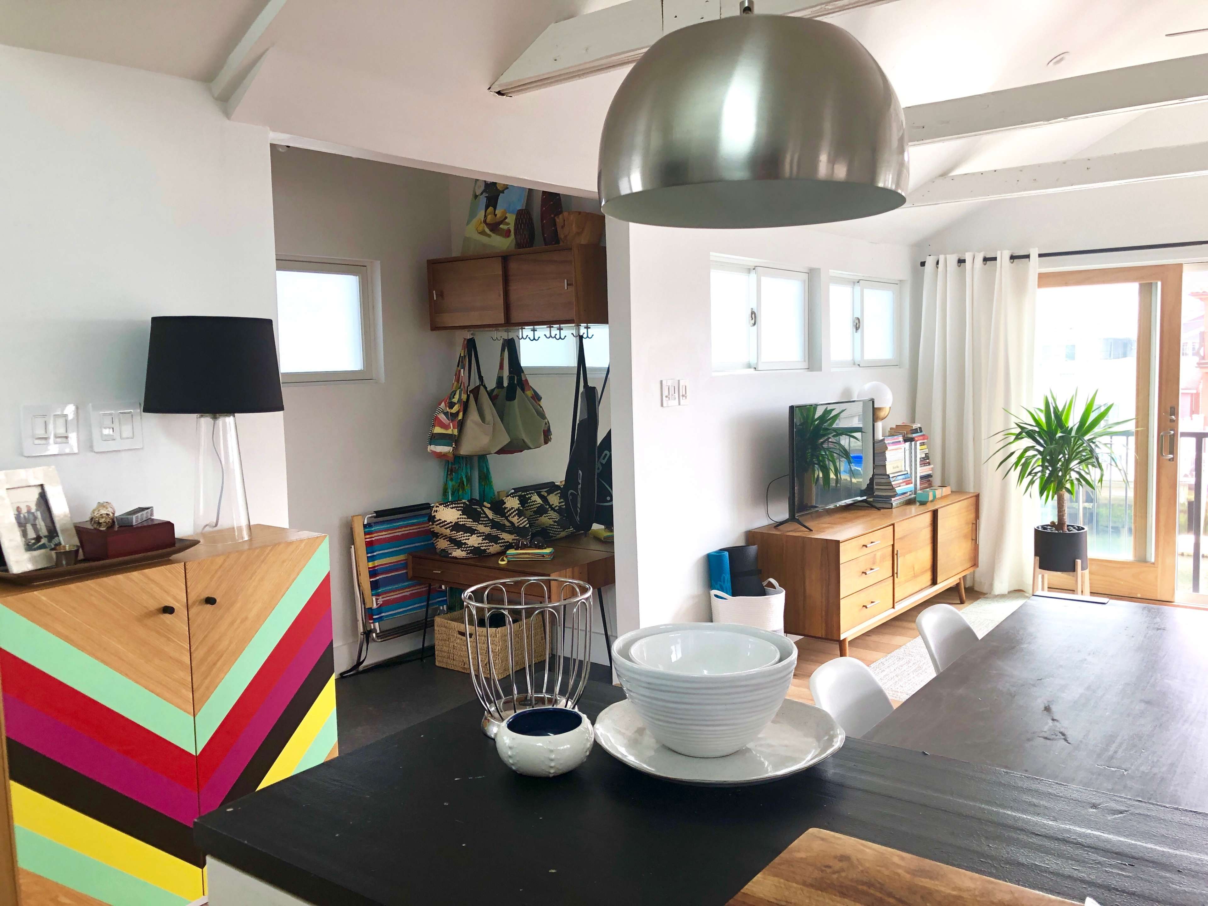 A modern living space features a dining area with a black table, colorful storage cabinet, and a well-lit corner with plants and shelves.