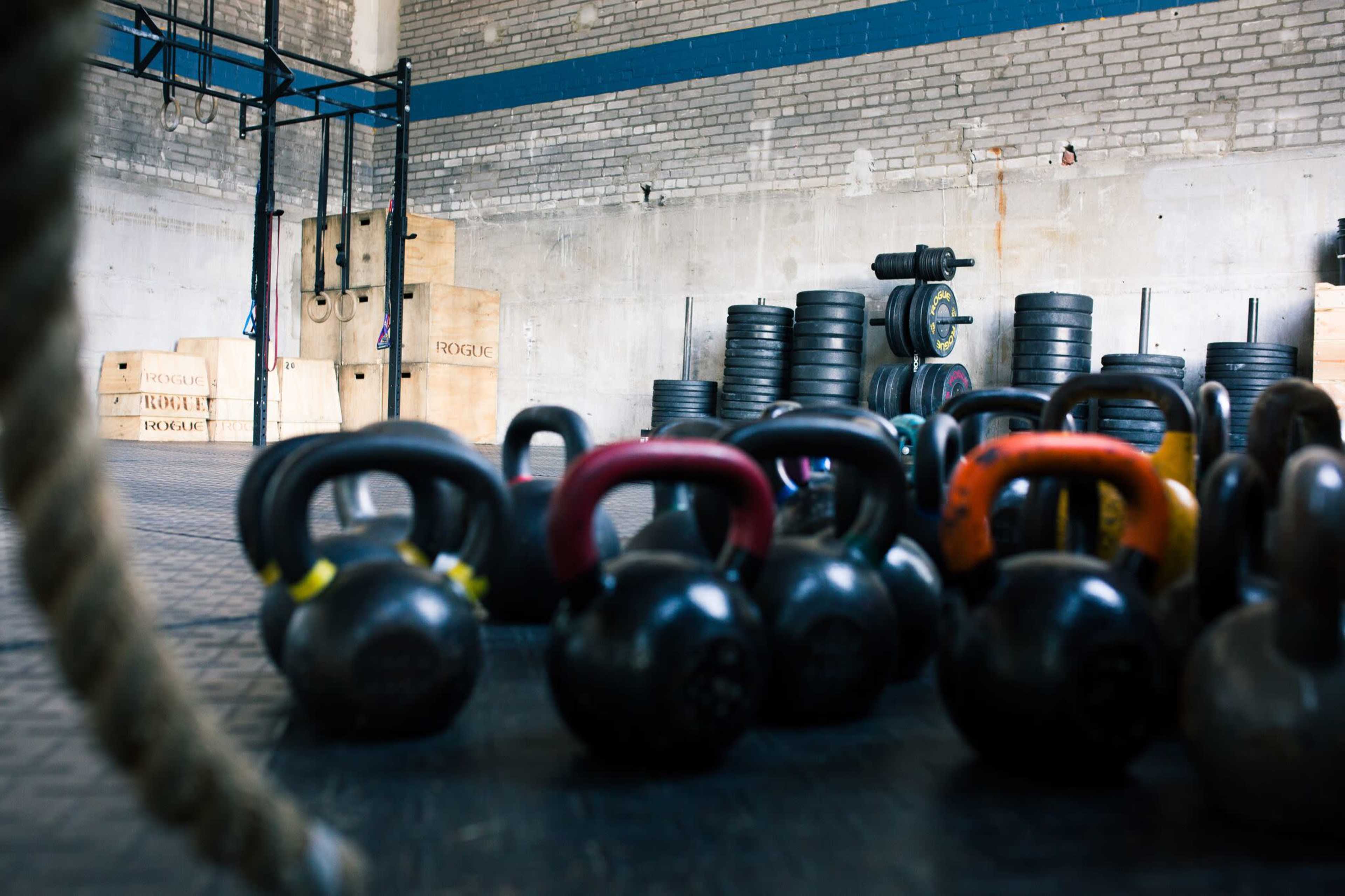 The image shows a gym interior with kettlebells in the foreground, stacked weight plates in the background, and a set of gymnastic rings hanging from the ceiling.