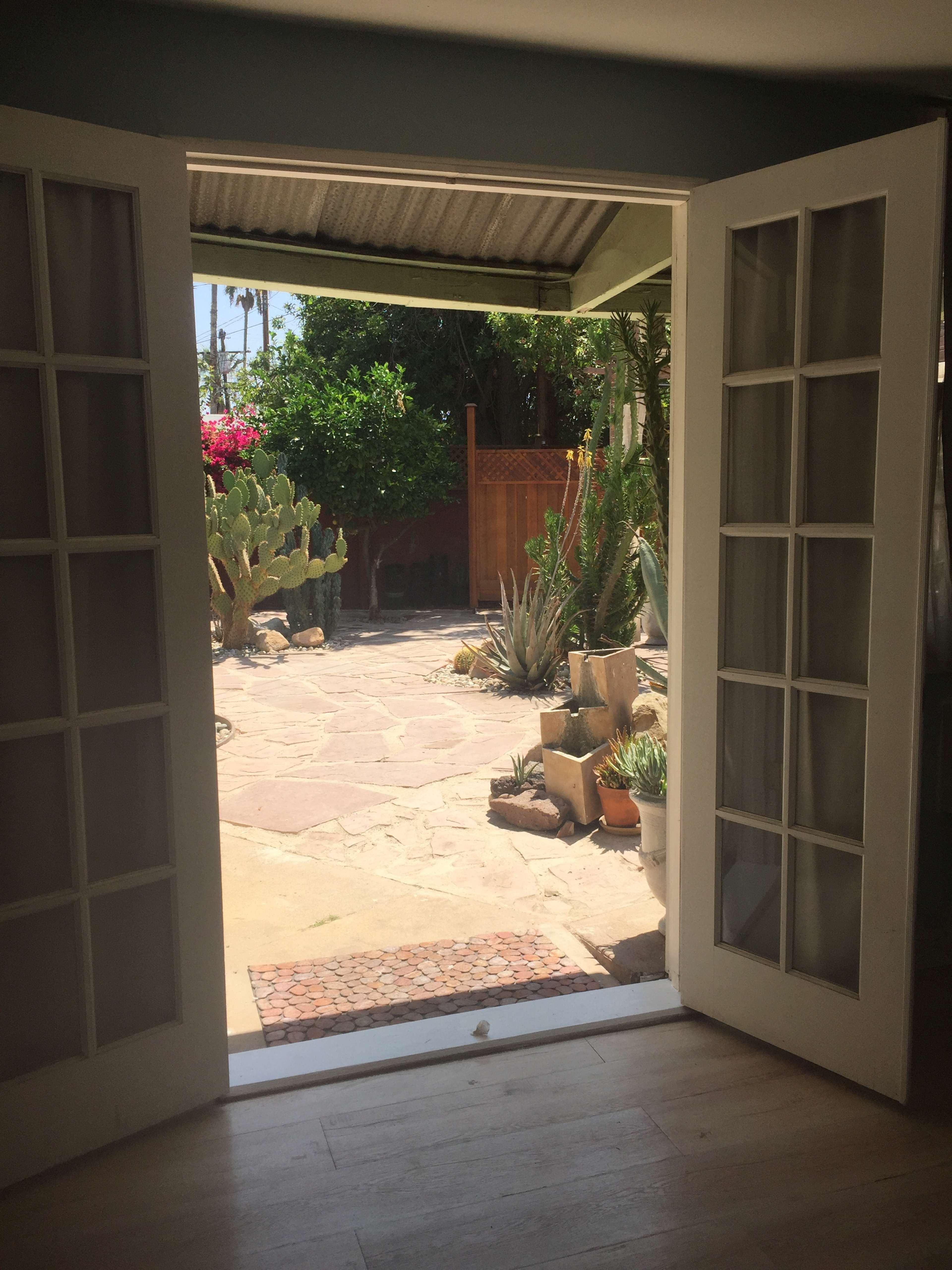 The image shows an open set of double doors leading to a stone patio surrounded by various plants and cacti.