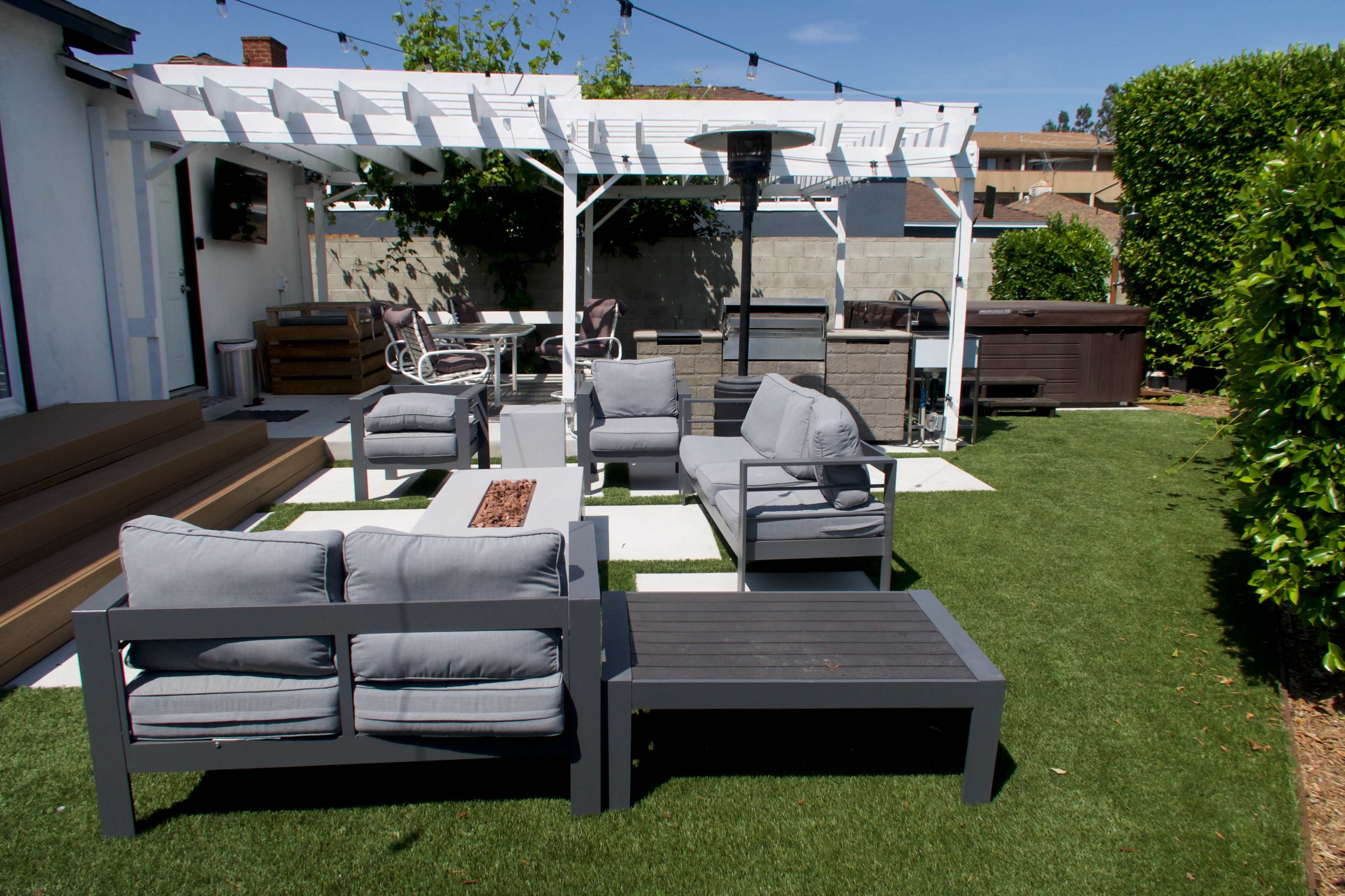 An outdoor seating area with gray furniture on a green lawn, surrounded by a pergola and a hot tub in the background.