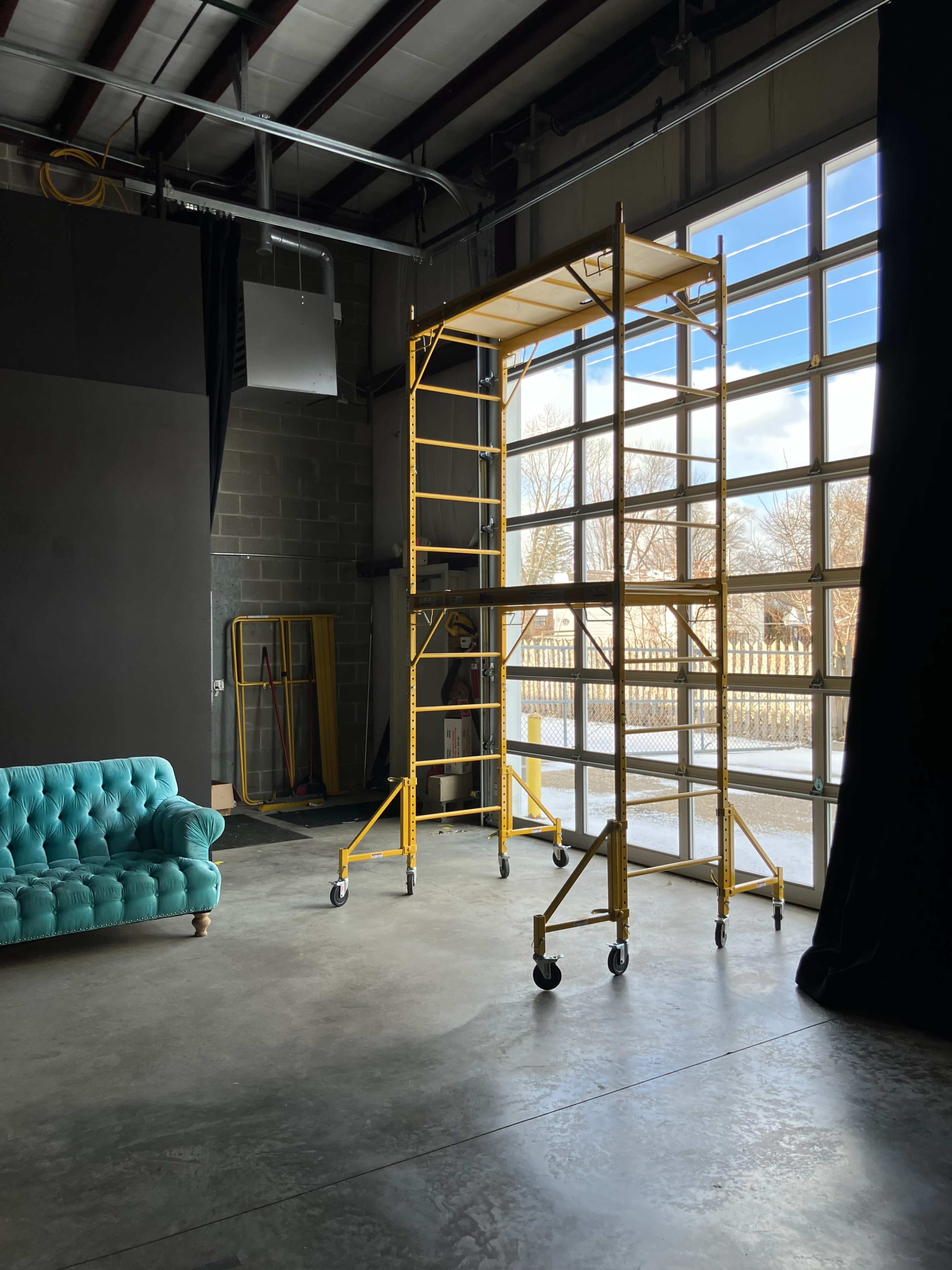 A turquoise couch is positioned beside a mobile scaffolding structure in front of a large window in an industrial space.