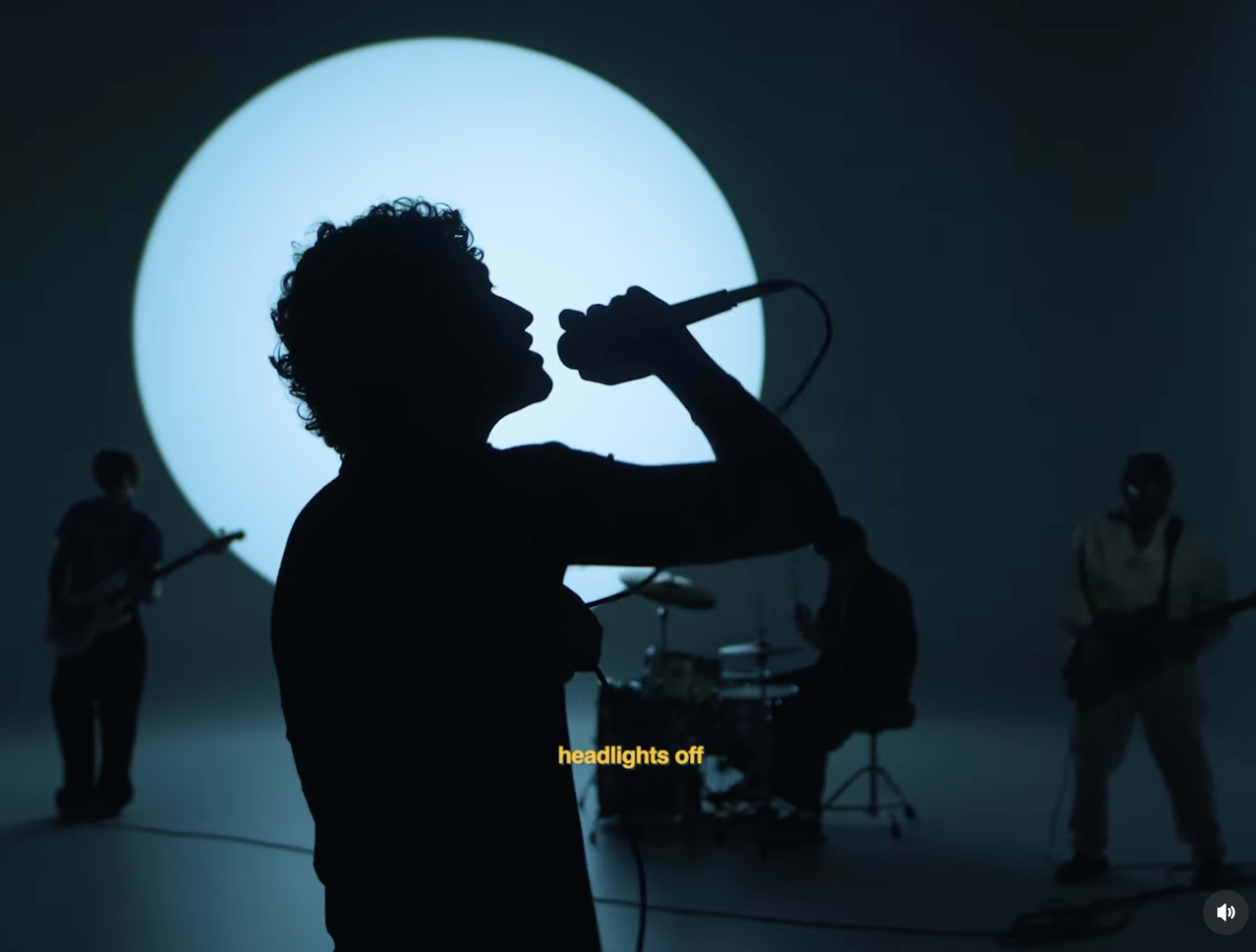 A silhouetted band performs in front of a large illuminated circle on a dark set.