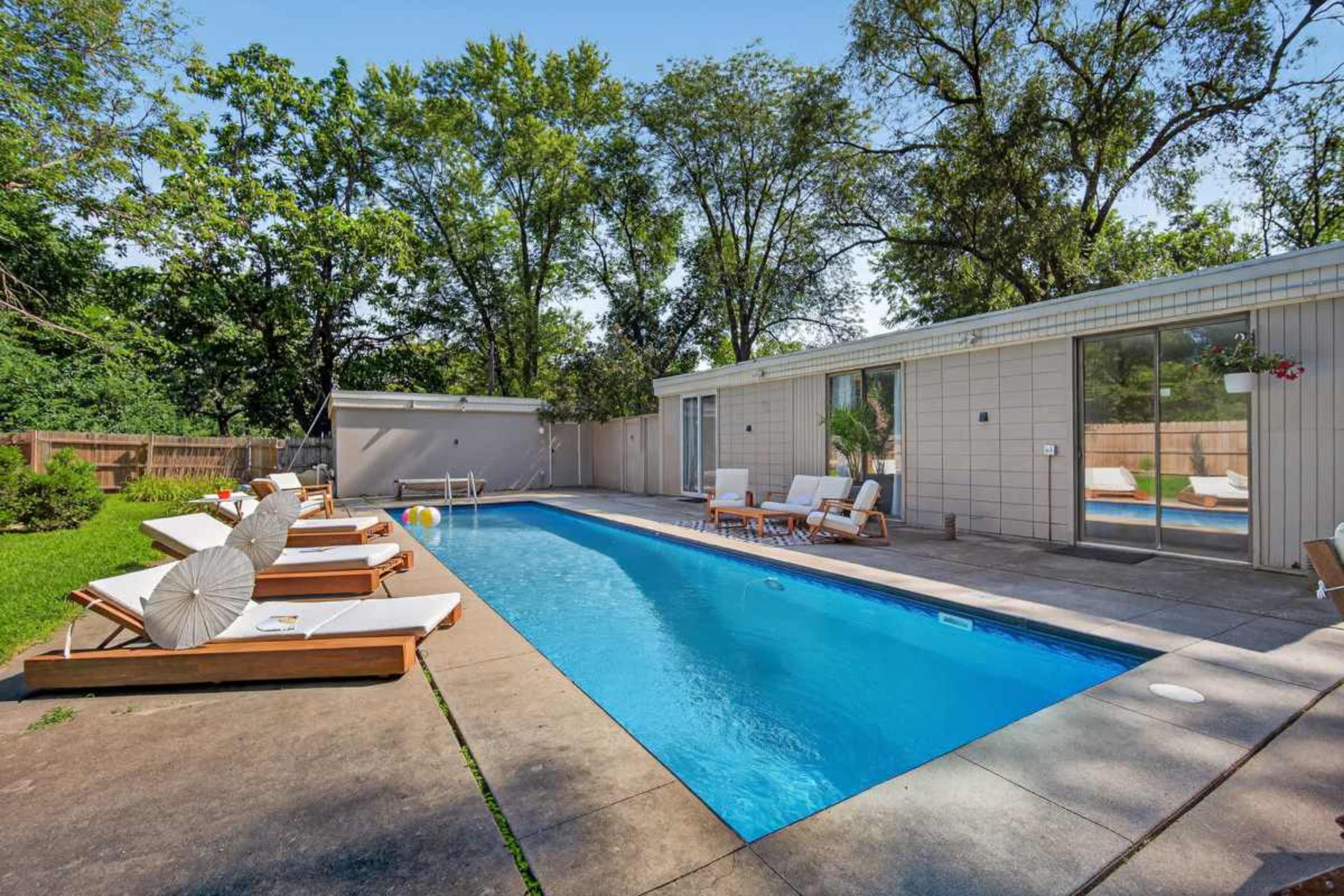 A rectangular swimming pool is surrounded by wooden lounge chairs and greenery, with a light-colored building visible in the background.
