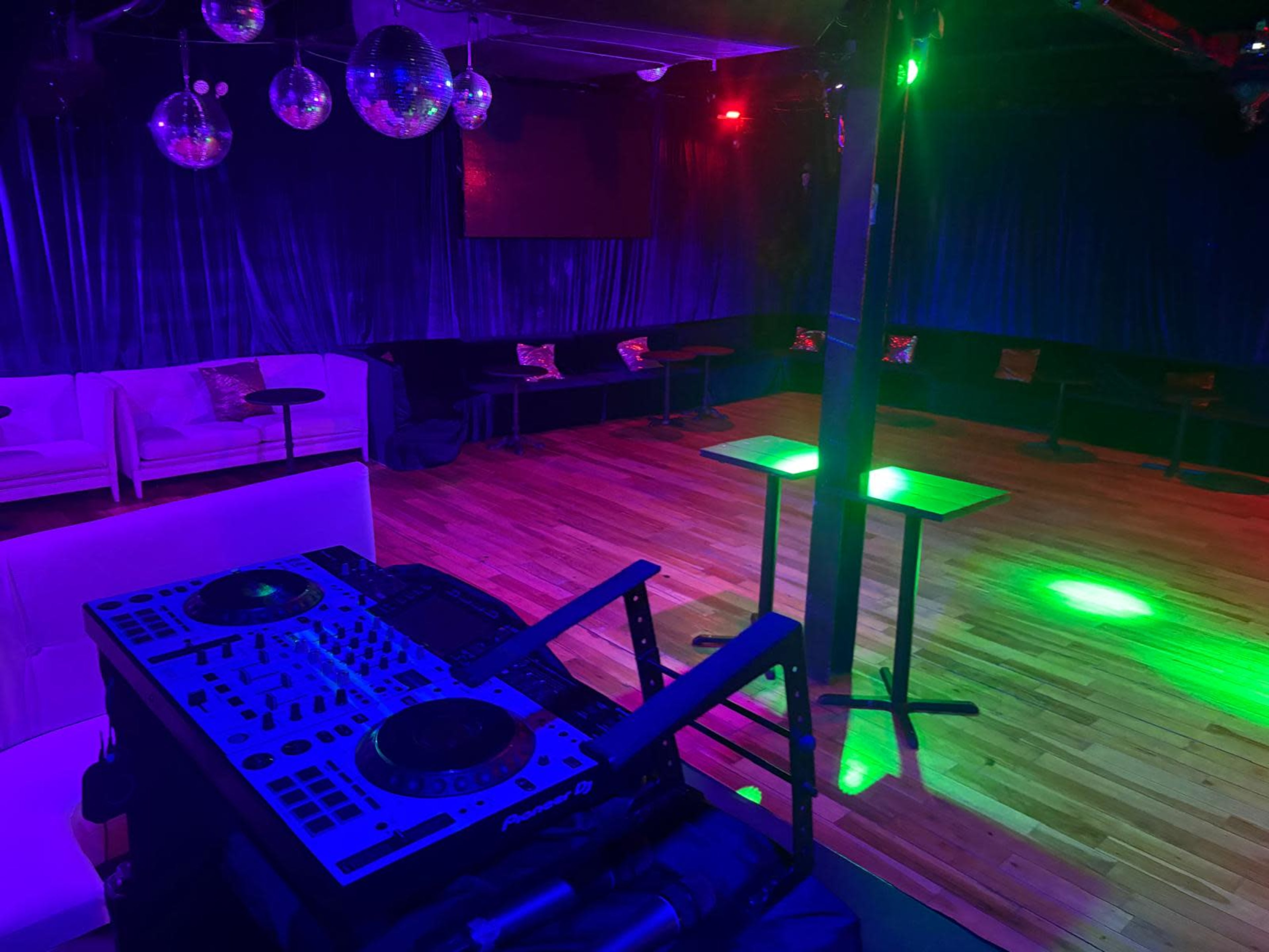 The image shows a dimly lit dance floor with a DJ setup, surrounded by seating areas and colorful lighting effects.