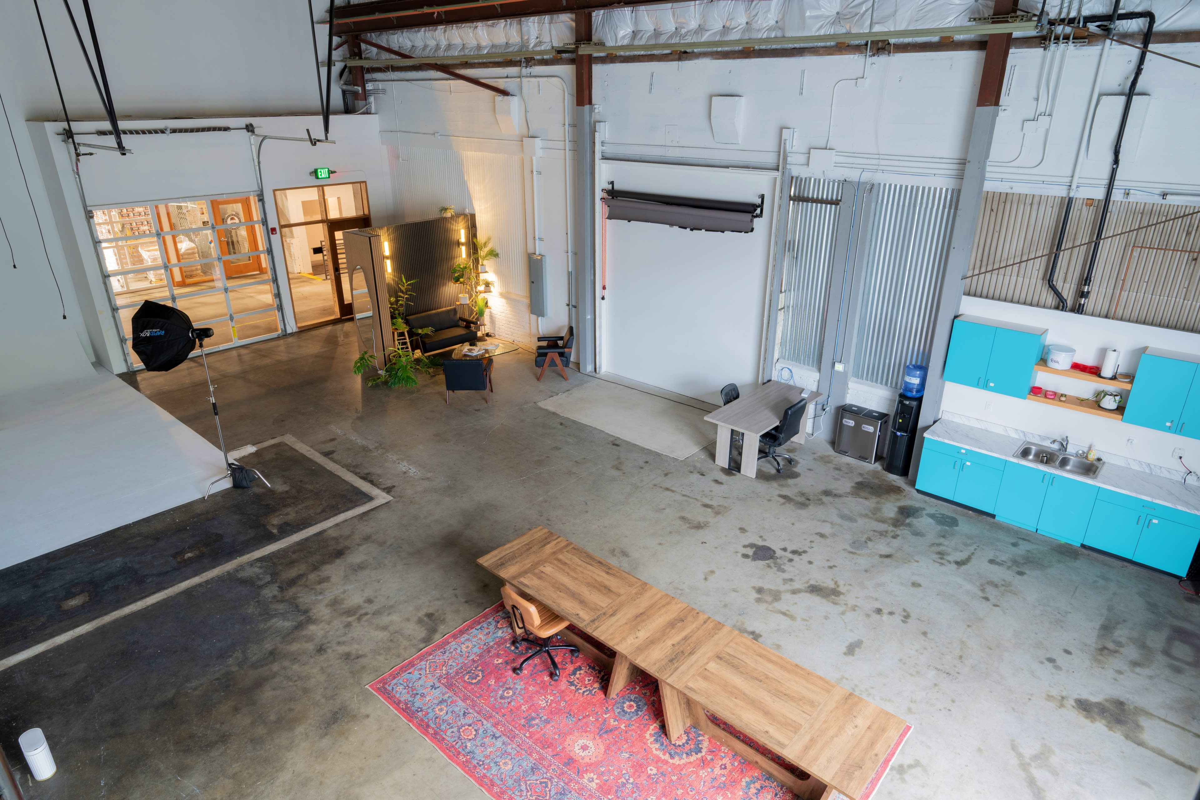 The image shows a spacious, well-lit industrial studio with a wooden table, an office chair, and a kitchenette with blue cabinets.