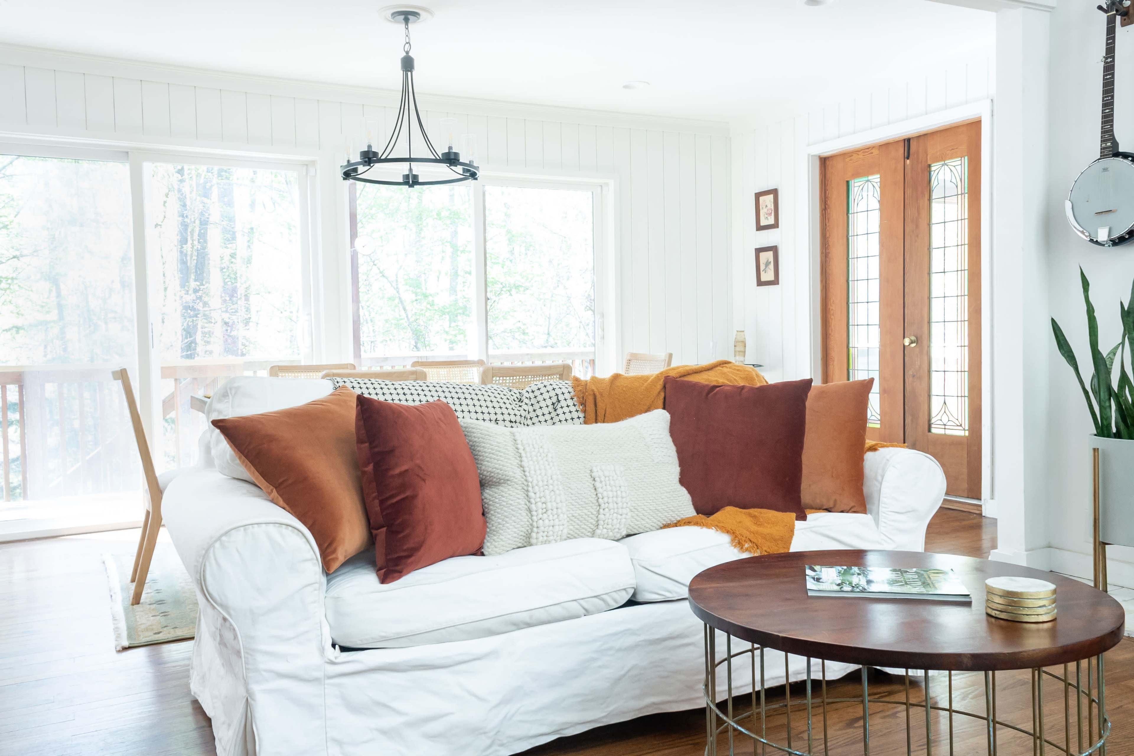 A white couch adorned with various textured pillows sits in a bright room with large windows and wooden doors.