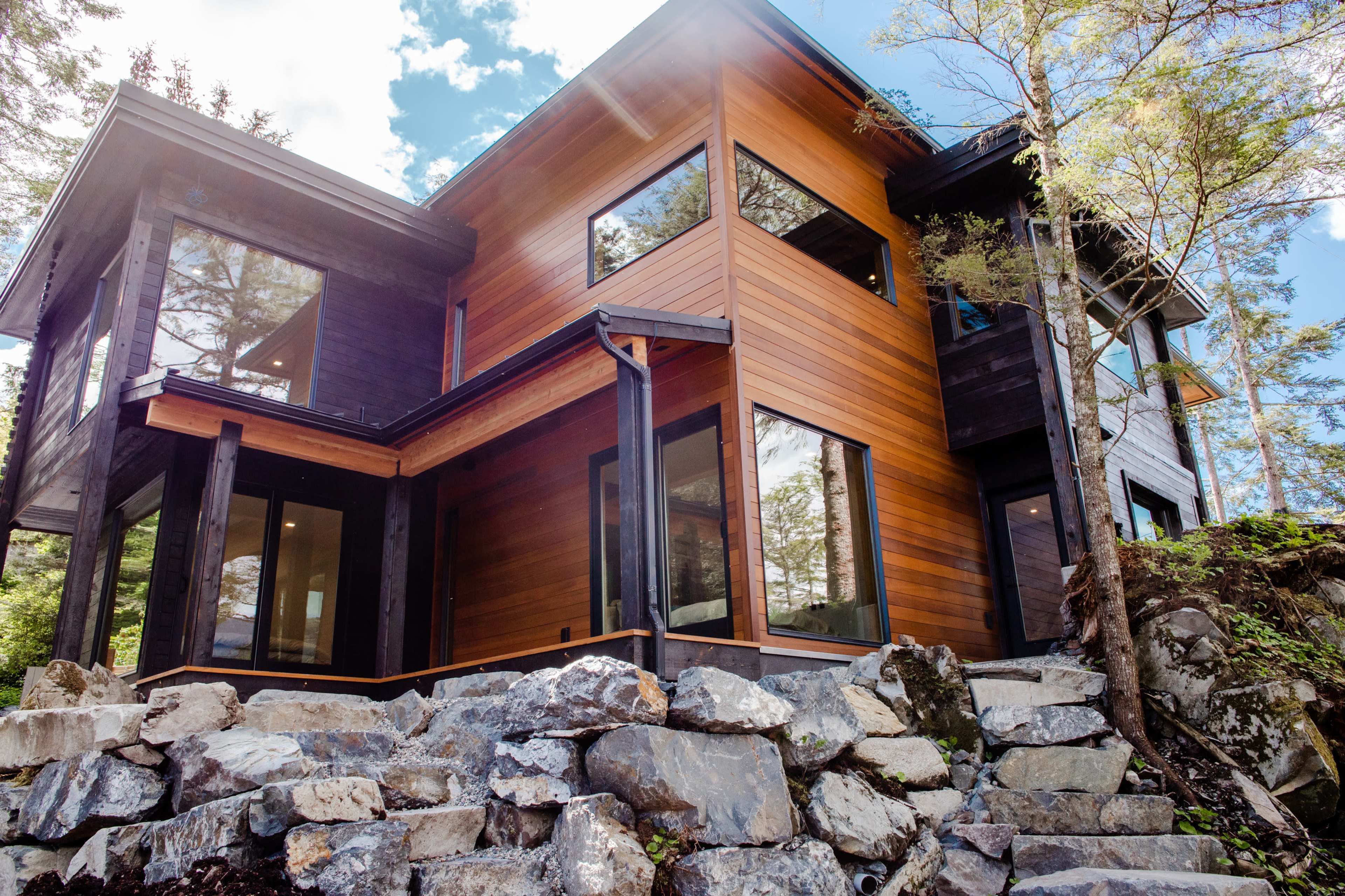 A modern, multi-level house with large windows and a mix of wood and stone materials is set among trees and rocky landscaping.