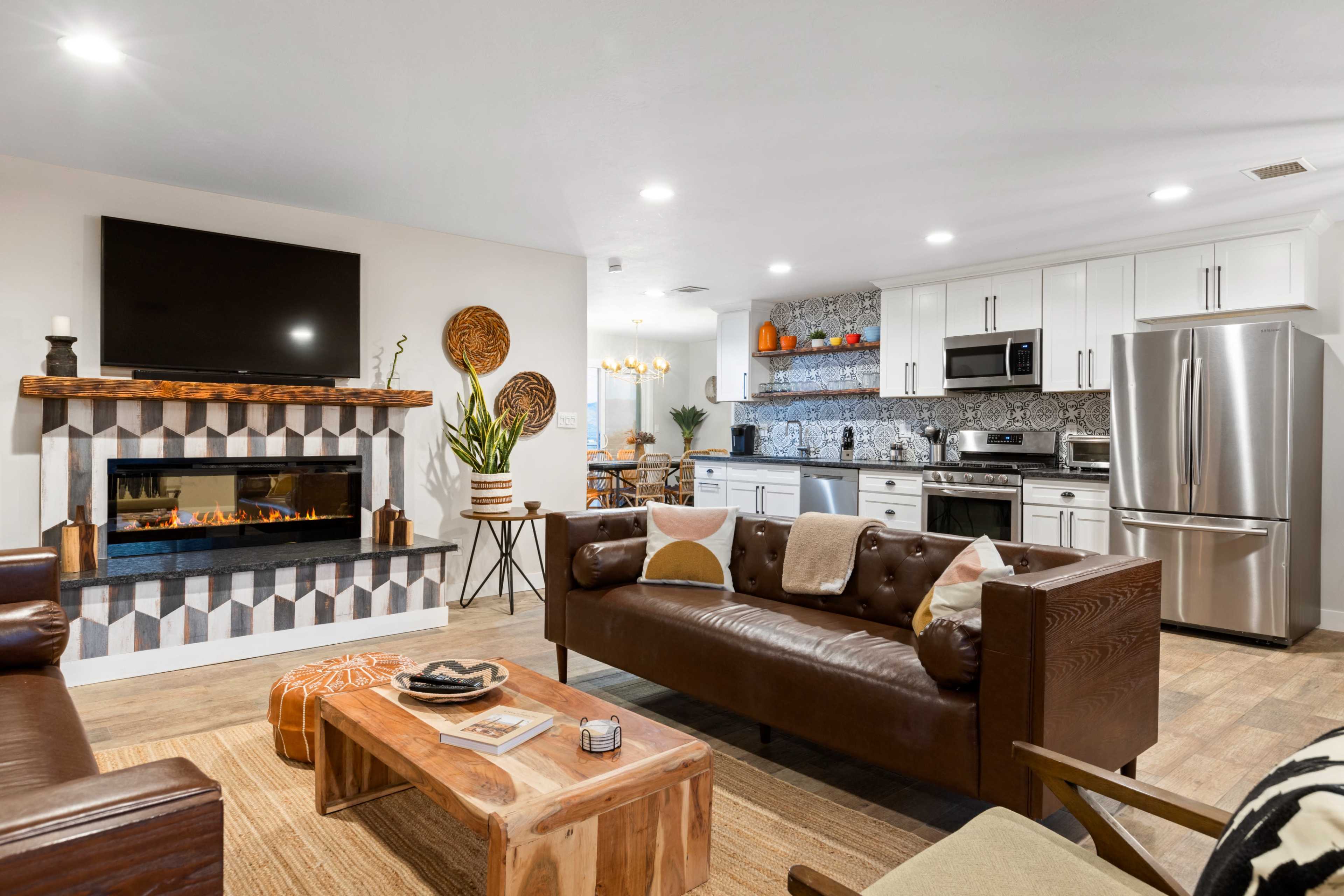 A modern living room features a brown leather sofa, a wooden coffee table, and a decorative fireplace, with an open kitchen visible in the background.