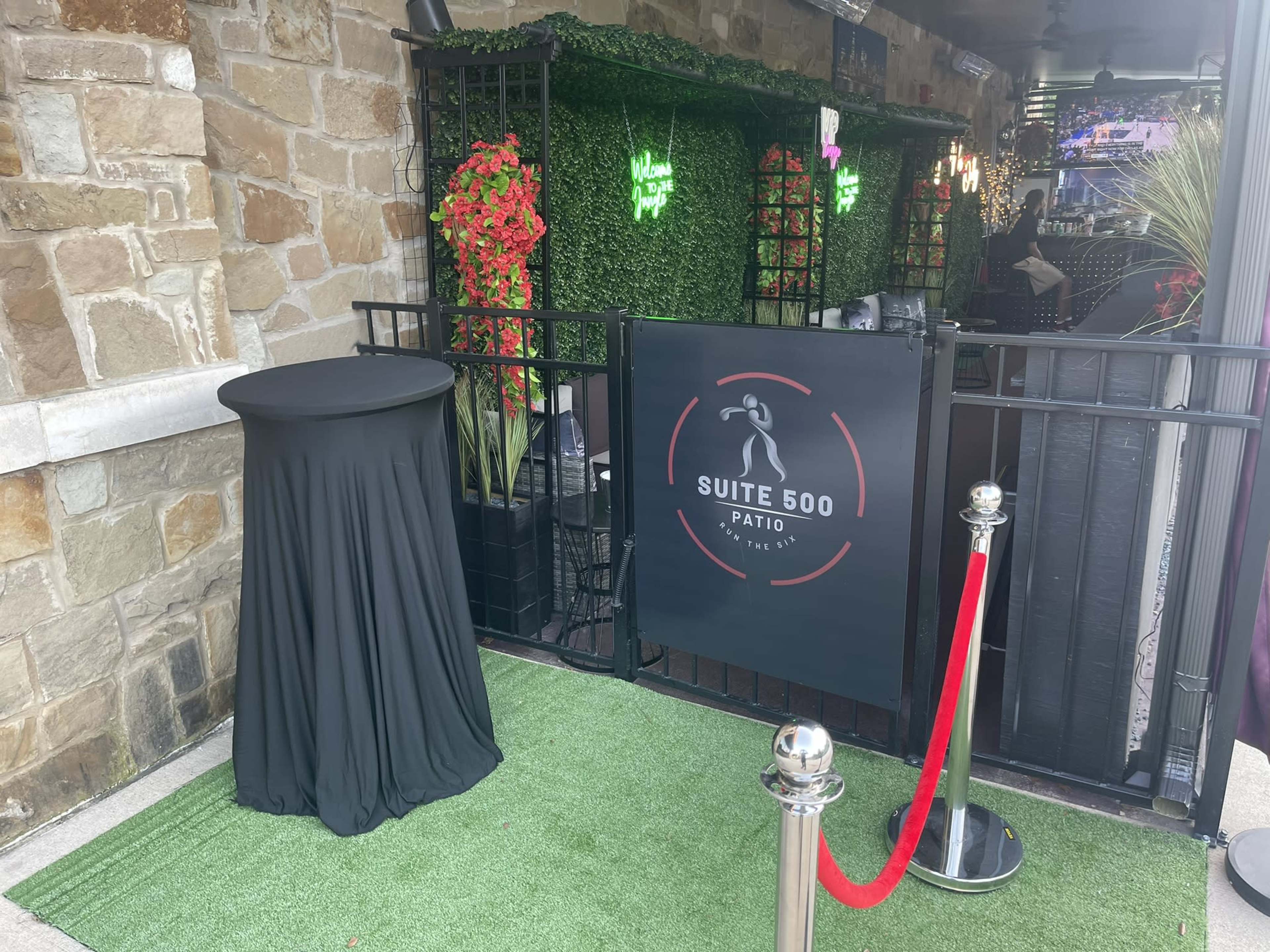 The entrance to Suite 500 features a black table draped in fabric and a red rope barrier leading to a green patio area adorned with plants and neon signs.