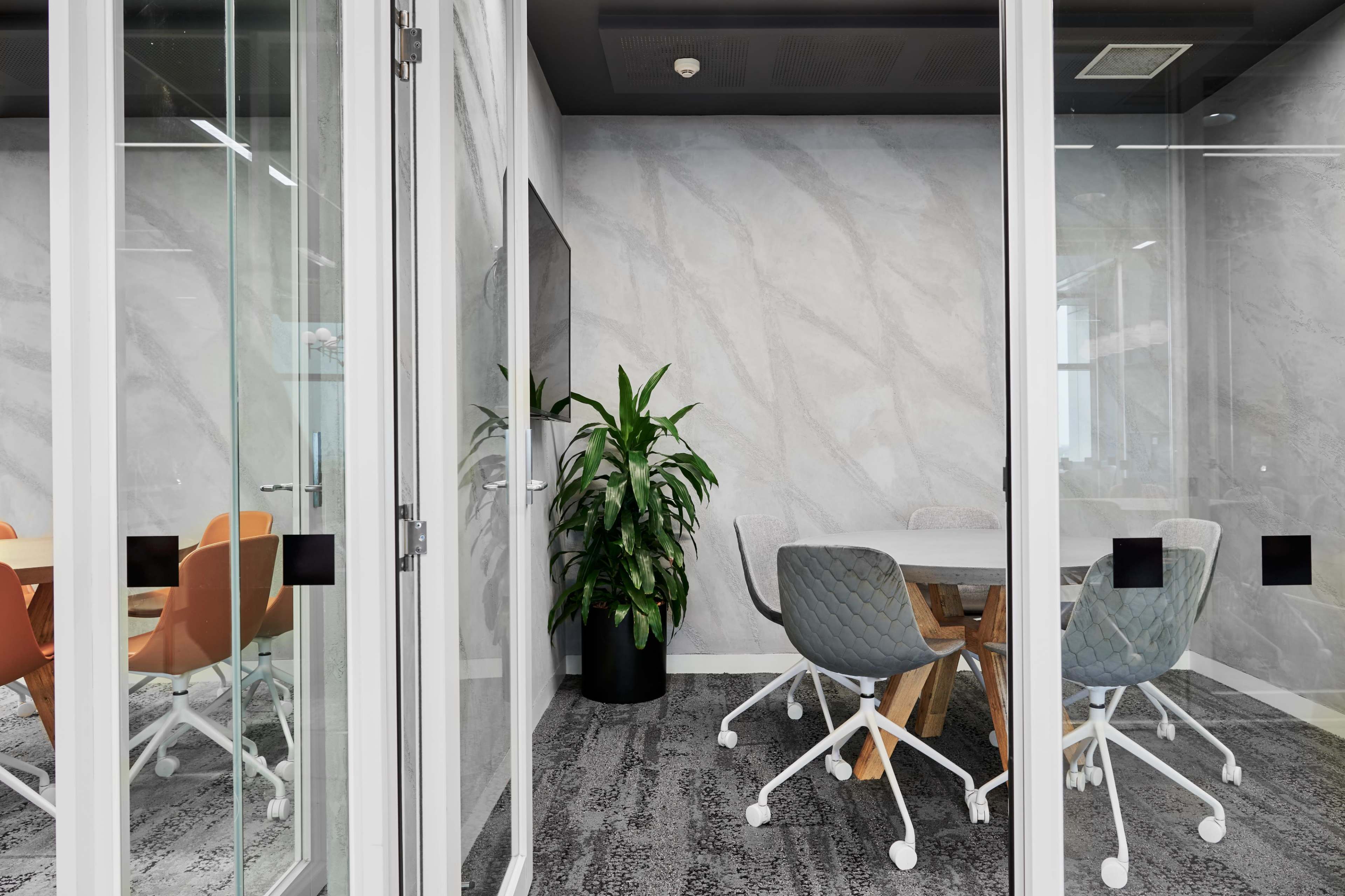 A conference room with a wooden table surrounded by gray and orange chairs, enclosed by glass walls, features a large plant in the corner.