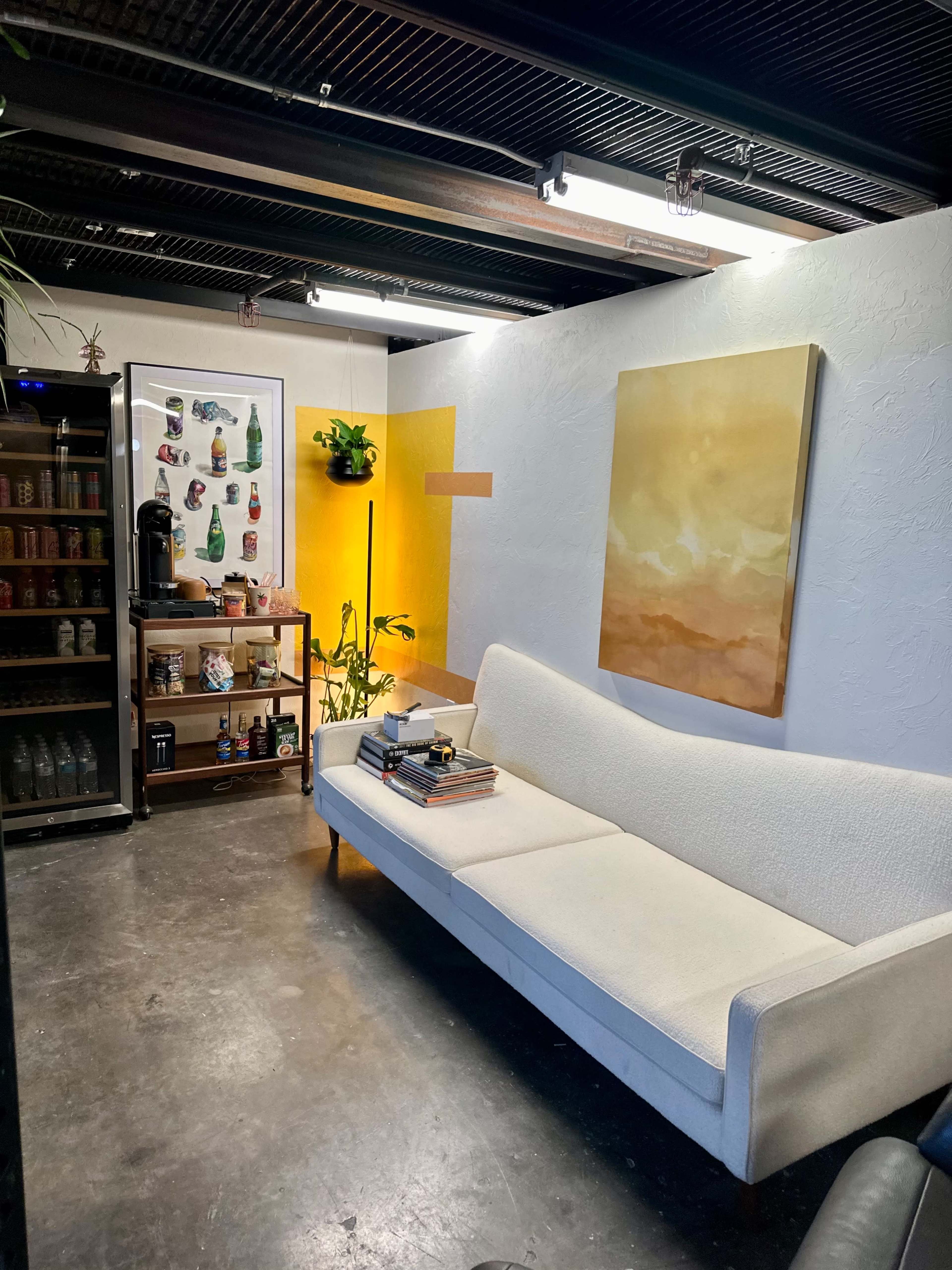 A cozy room with a white sofa, a wooden shelf filled with books and decor, a coffee station, and a wall adorned with art and a plant.