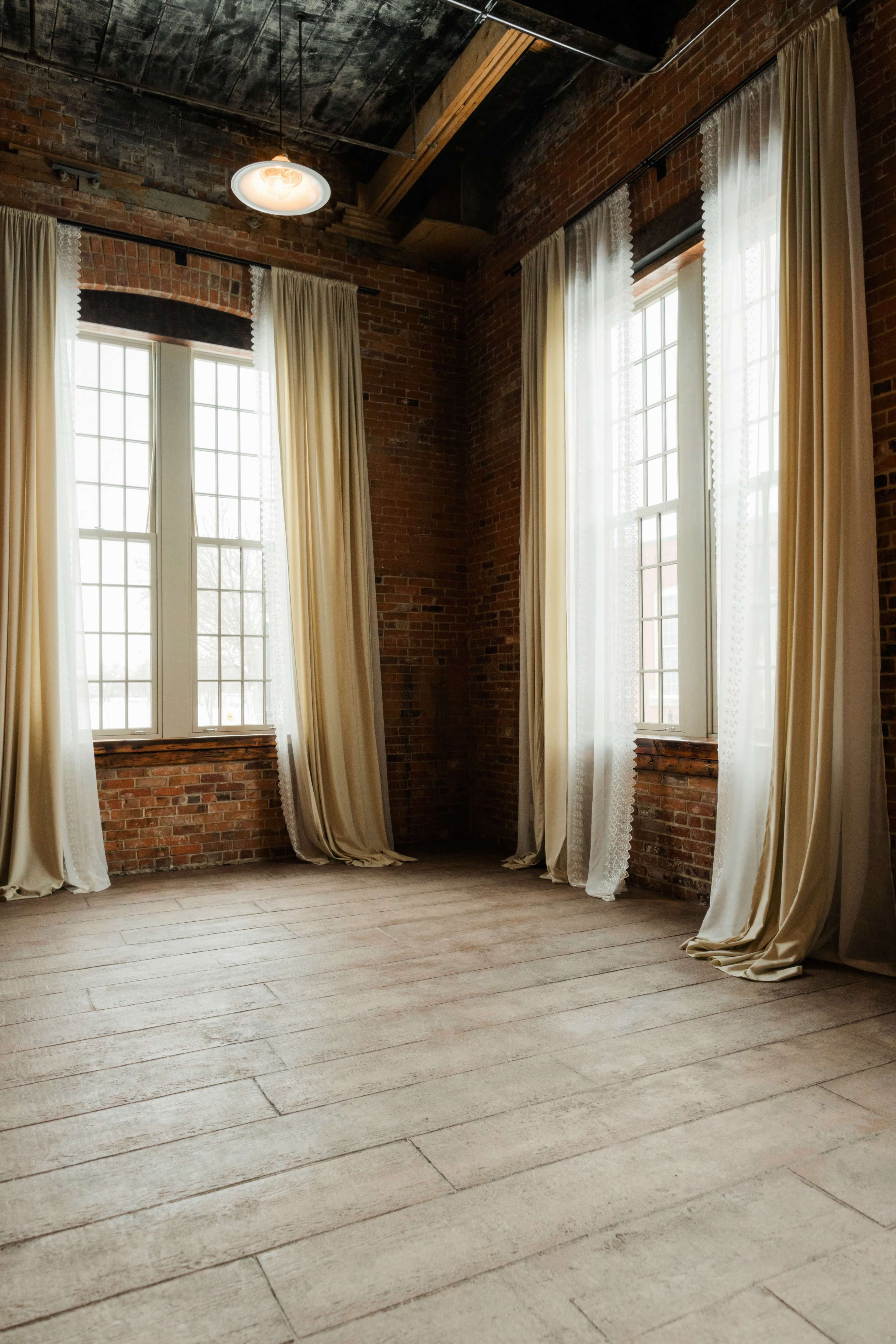 A spacious indoor area with large windows and light curtains, featuring exposed brick walls and wooden flooring.