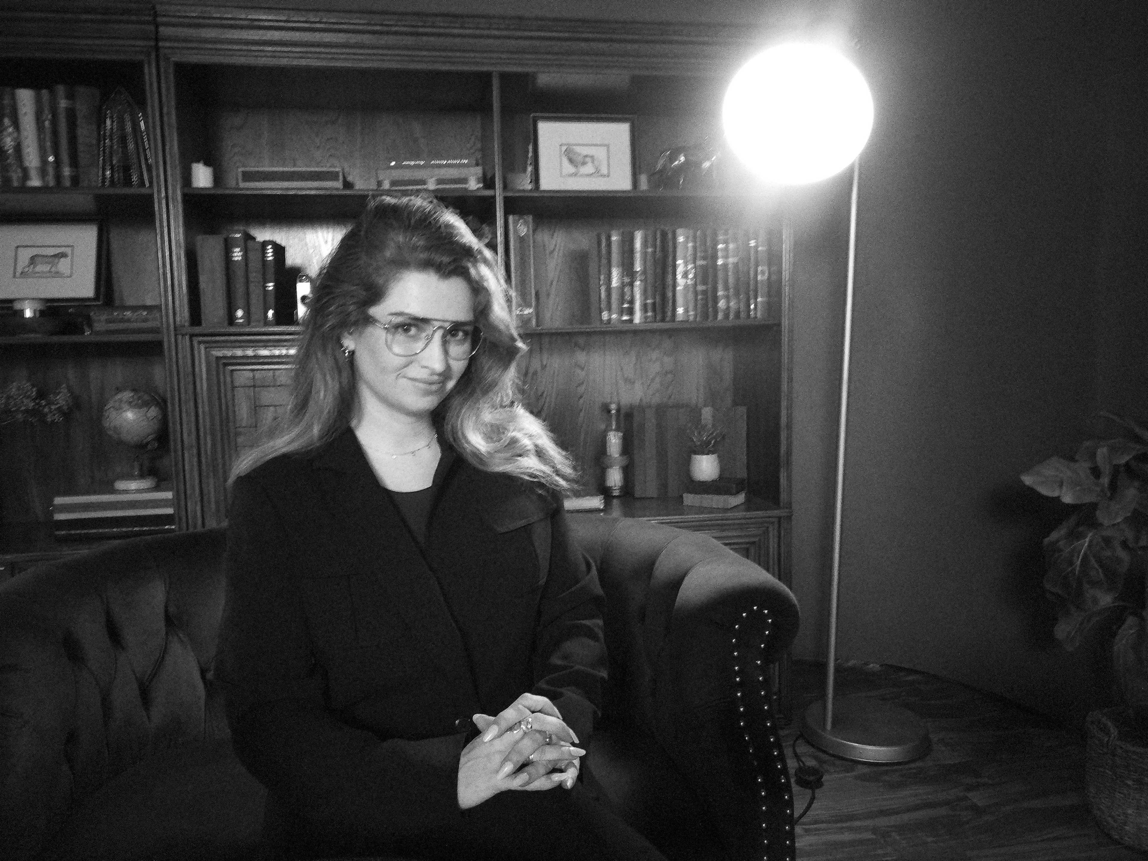 A woman with glasses sits on a dark couch in front of a wooden bookshelf and a floor lamp.