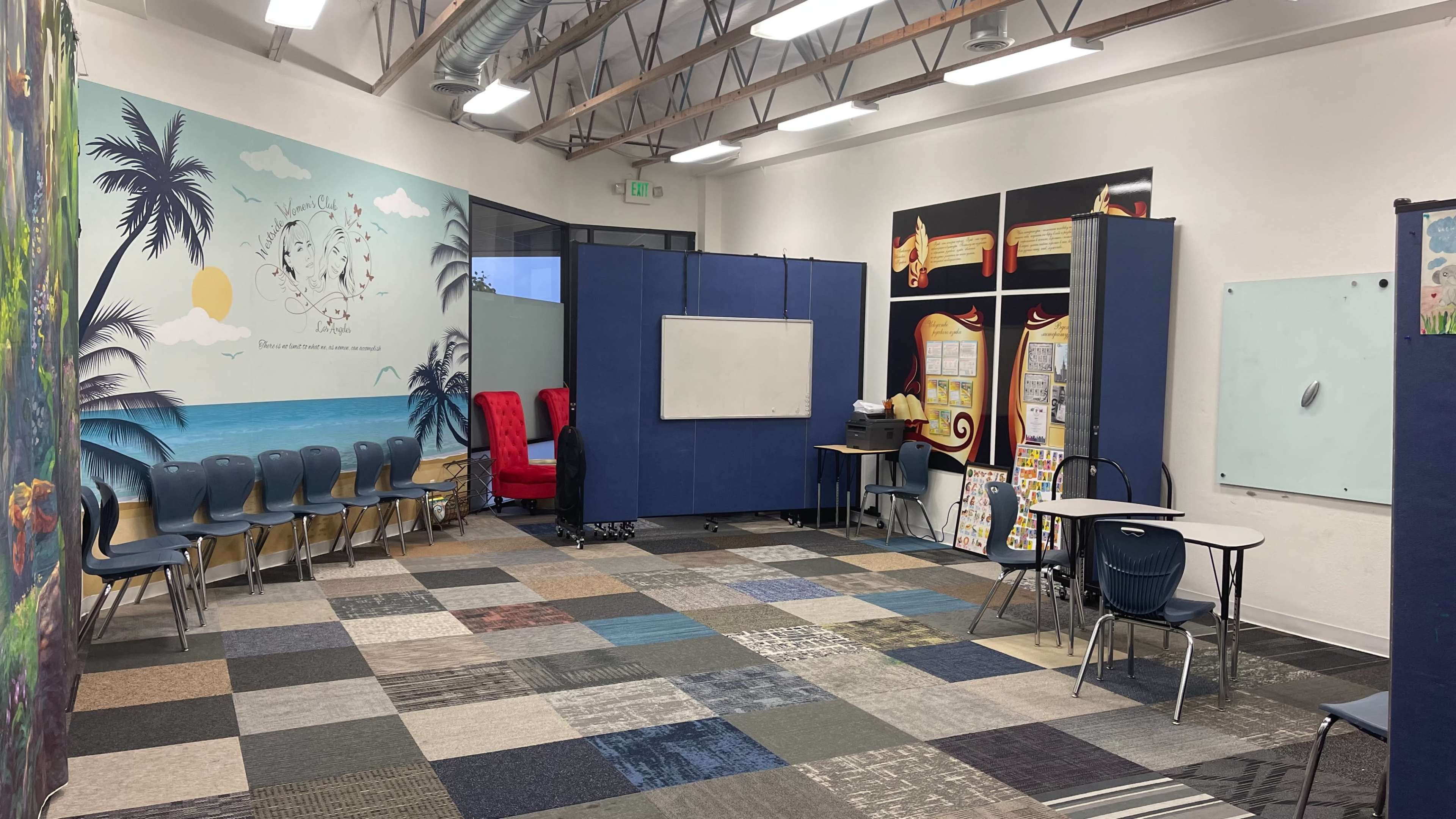 A classroom with blue partitions, colorful carpet tiles, and seating arrangements, including two red chairs and a table with various educational materials.