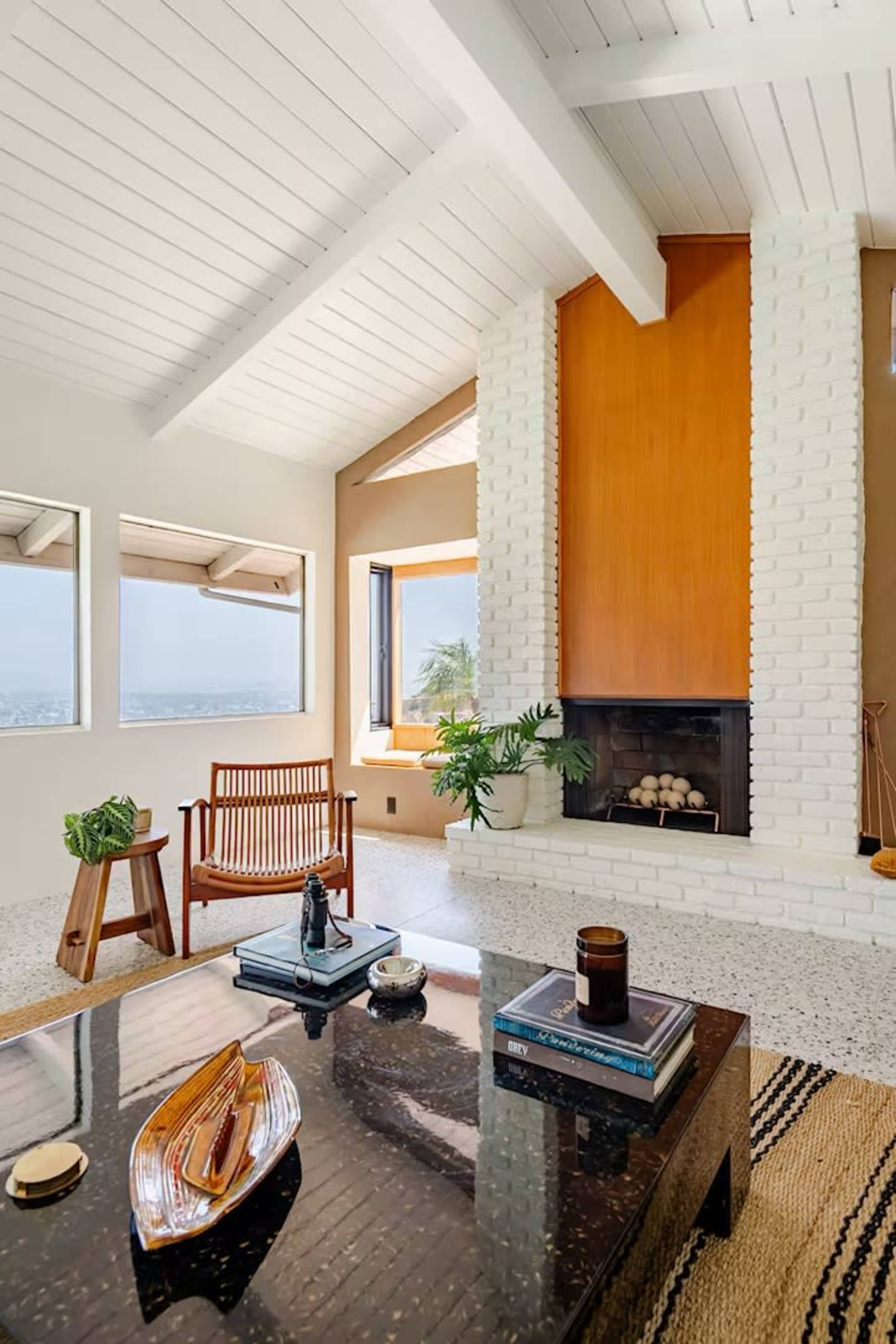 The image features a modern living room with a brick fireplace, a large window offering a view, and a mix of wooden and stone furniture.