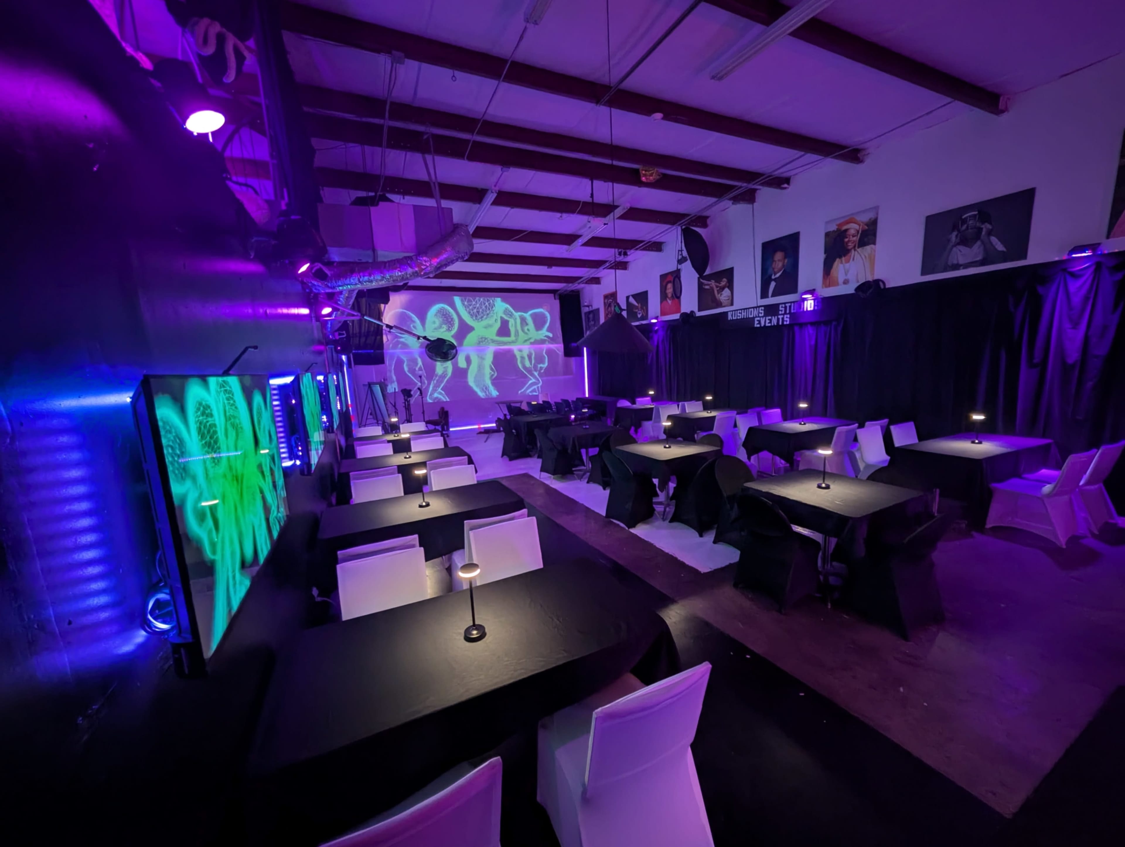 The image shows a dimly lit event space with black and white tables, illuminated by purple and green lighting, and featuring a projection on the wall.