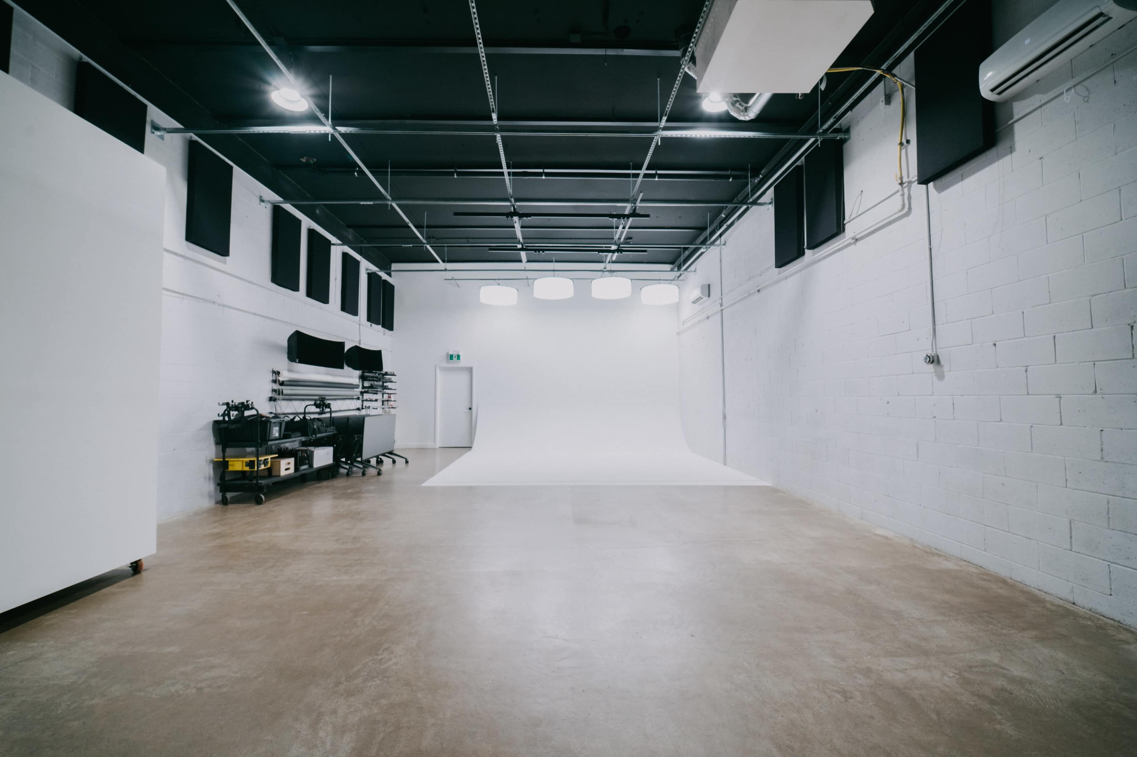 A large, empty photography studio features a plain white backdrop and concrete flooring, with equipment storage along one side.