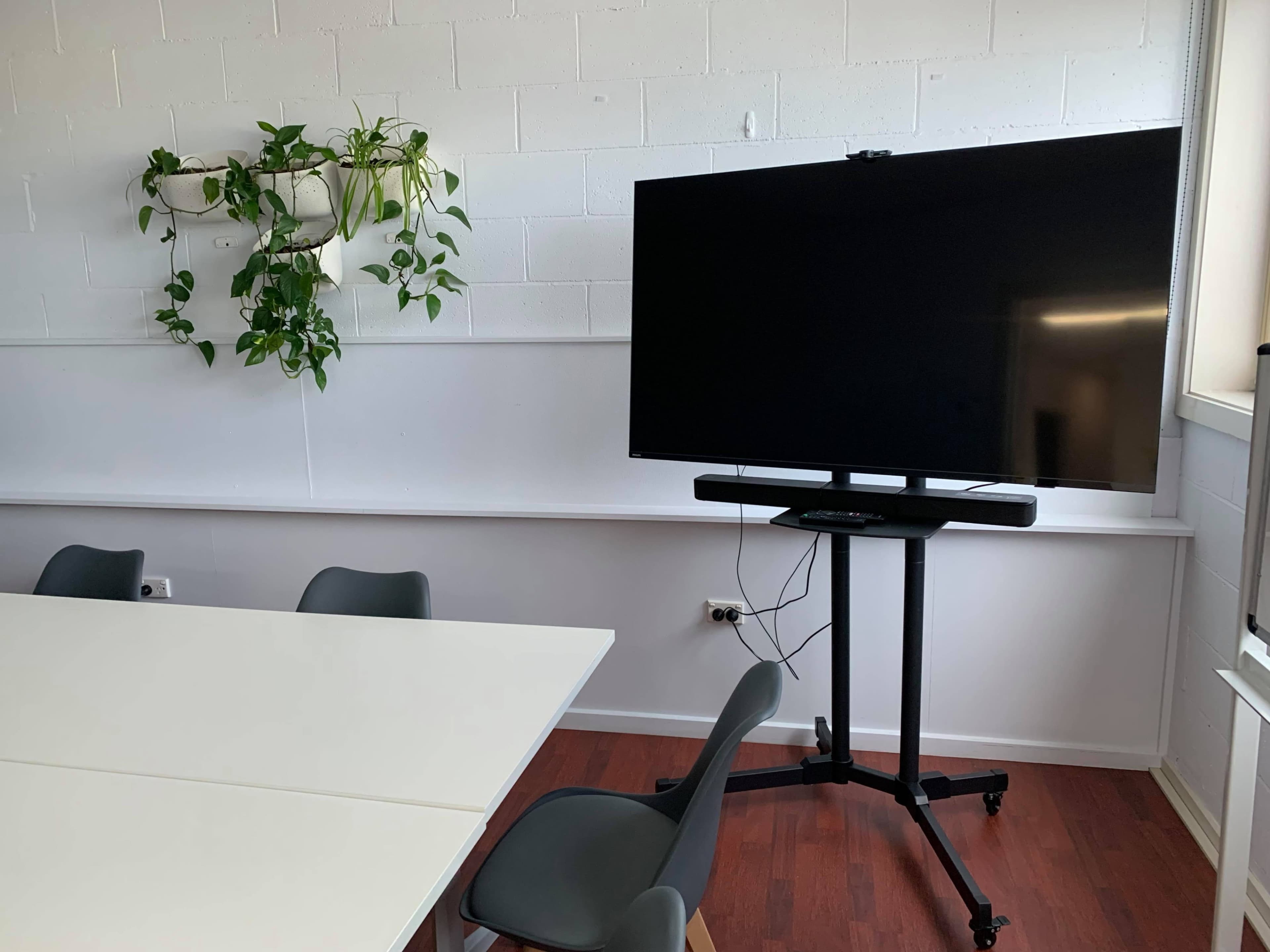 A modern meeting room features a large flat-screen television on a movable stand next to a table and green plants on the wall.