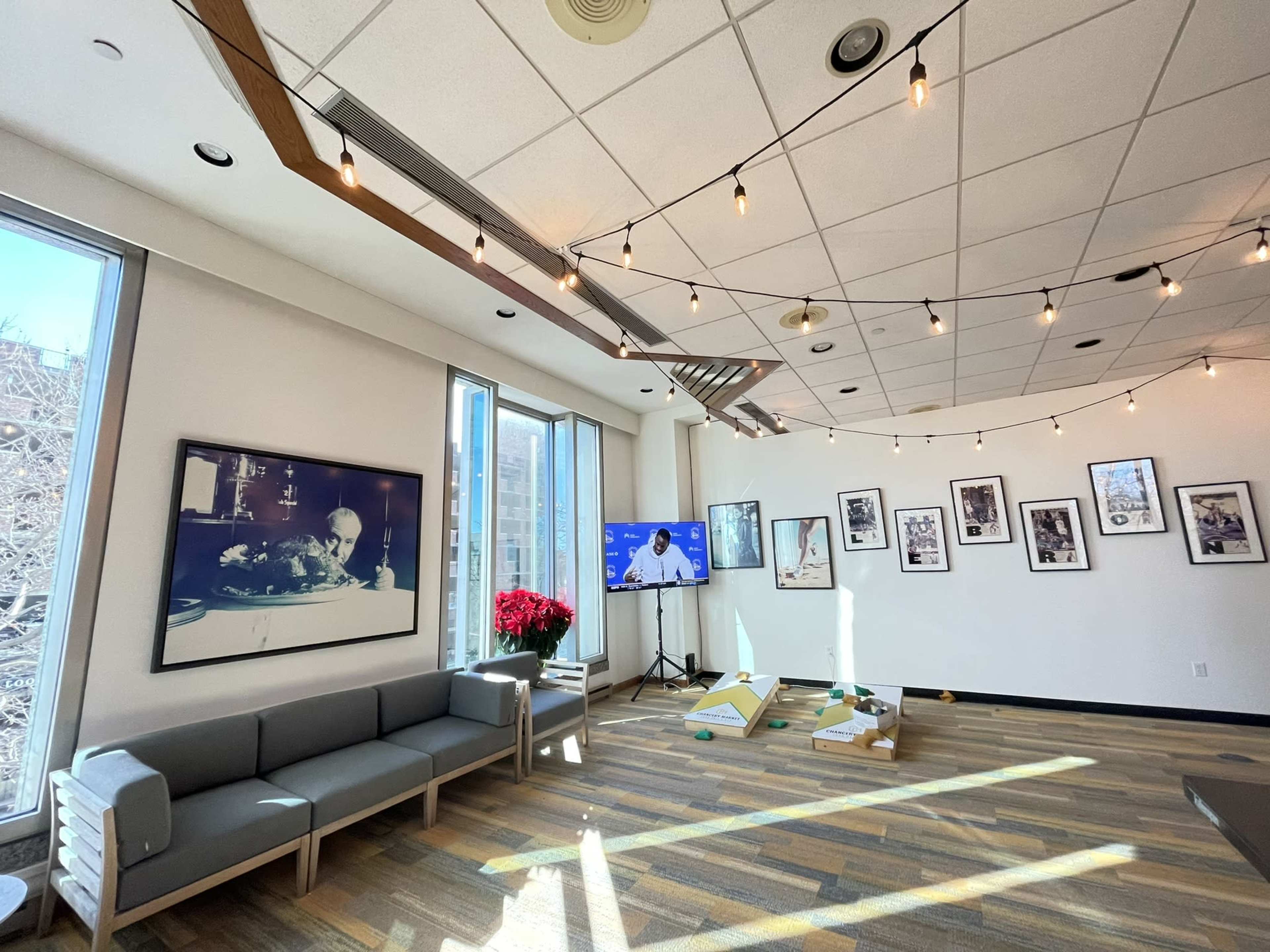 A well-lit room with large windows, a seating area with a gray couch, a television displaying a presentation, and framed photographs on the walls.