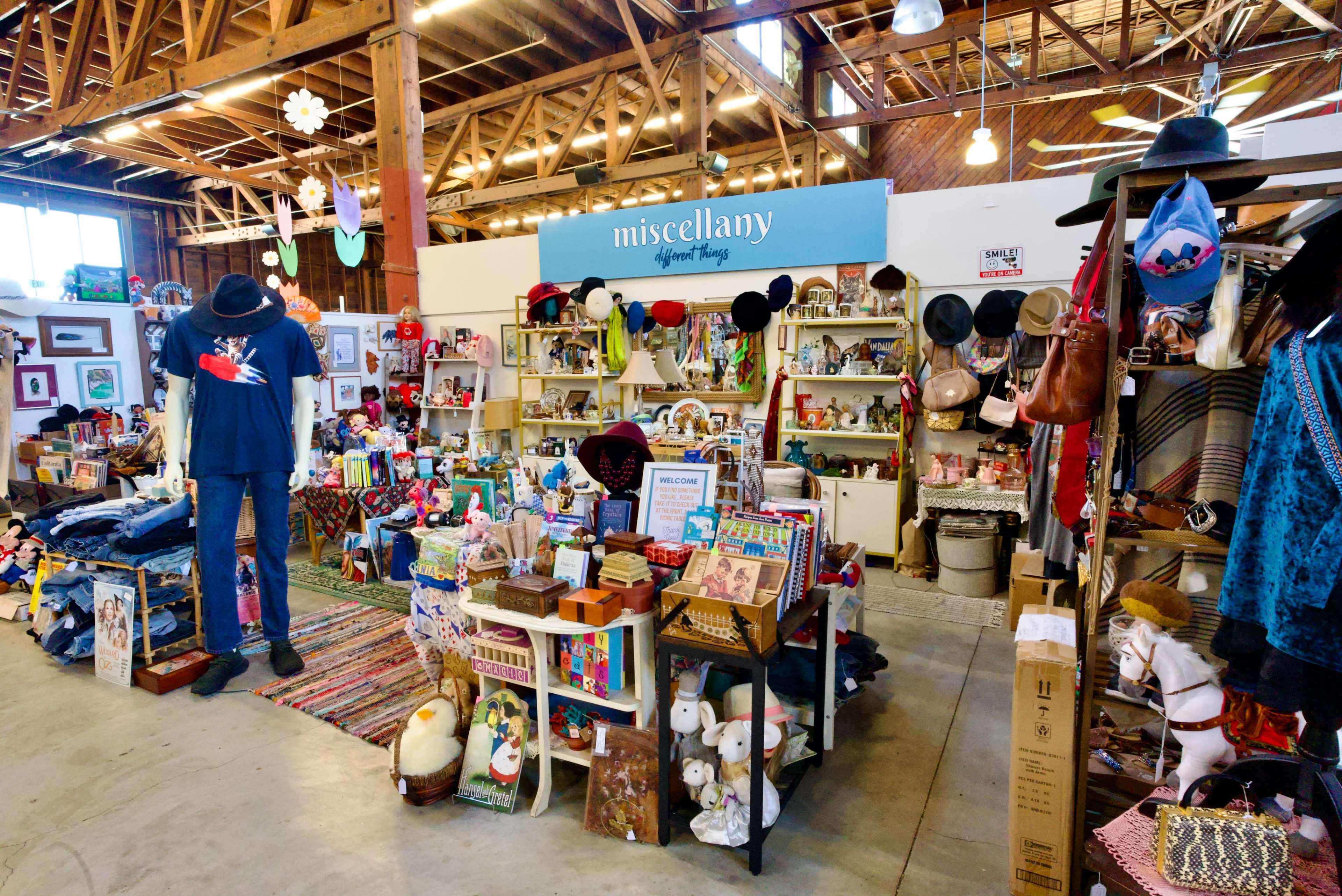 The image shows a vibrant indoor market space featuring a variety of colorful merchandise, including home decor, toys, and clothing, all displayed on shelves and tables.