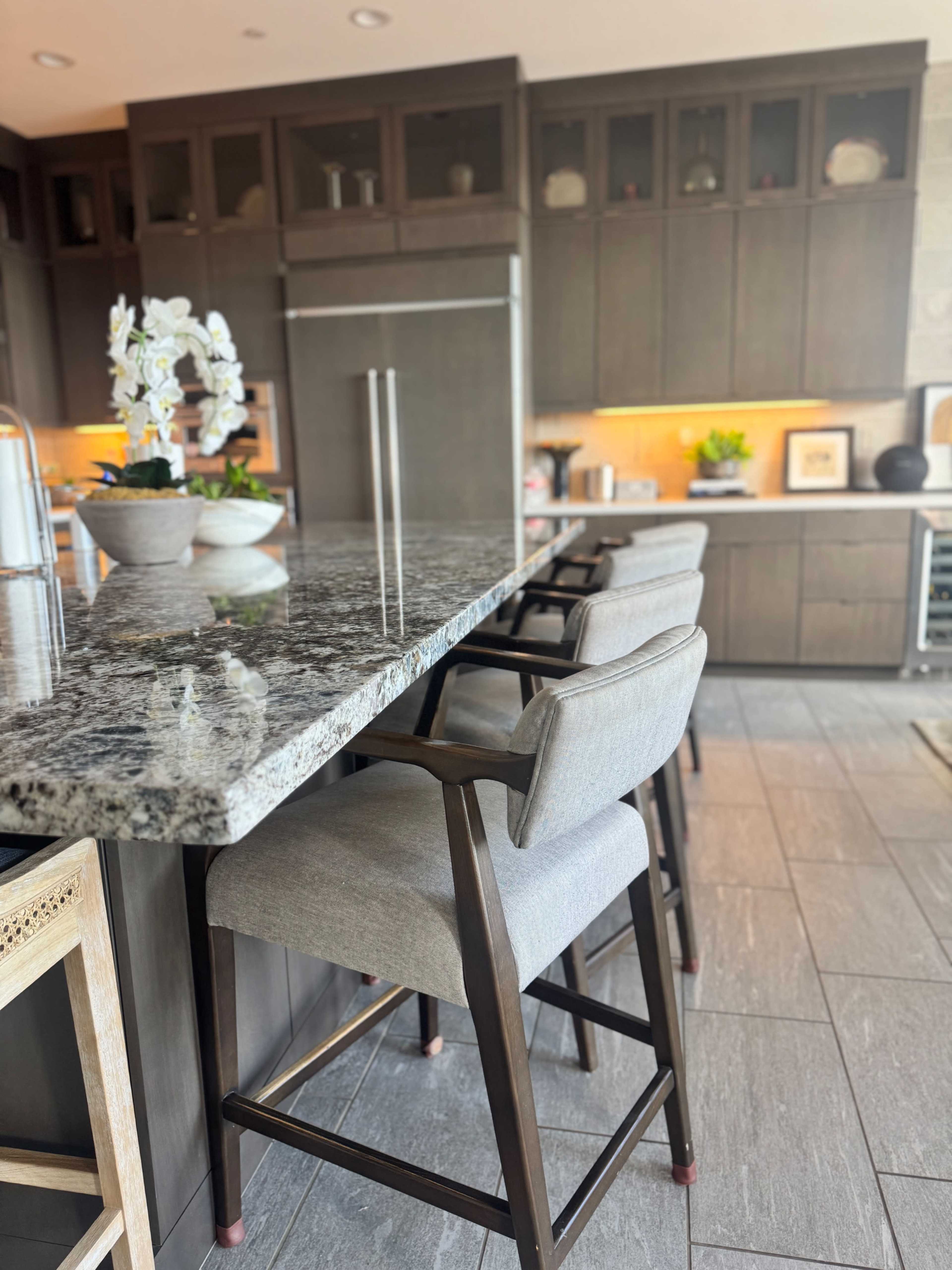 A modern kitchen features a granite countertop bar with three upholstered stools and dark wood cabinetry in the background.