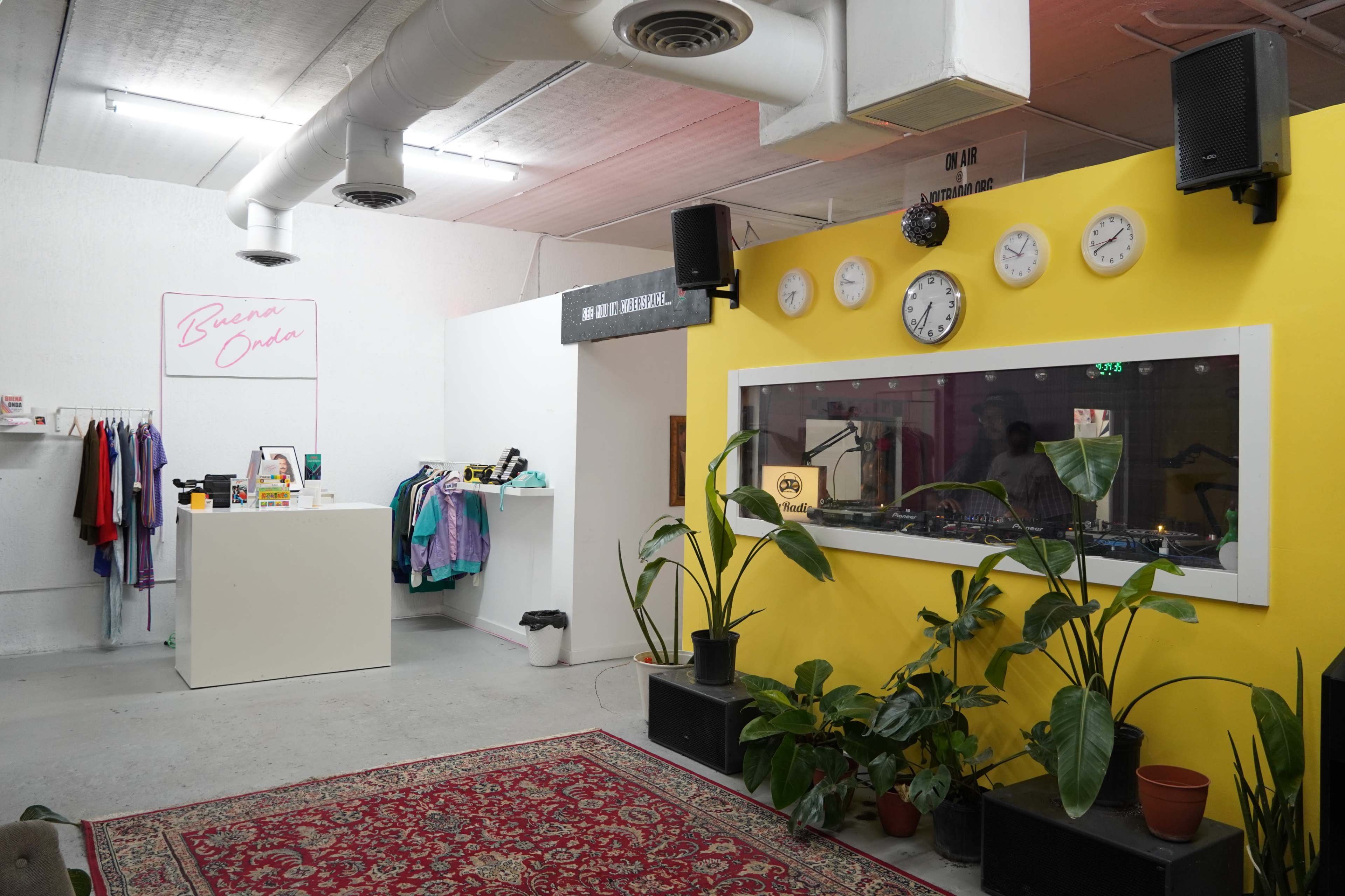 A clothing store interior with a bright yellow wall featuring multiple clocks, a display counter, and various plants on a dark platform.