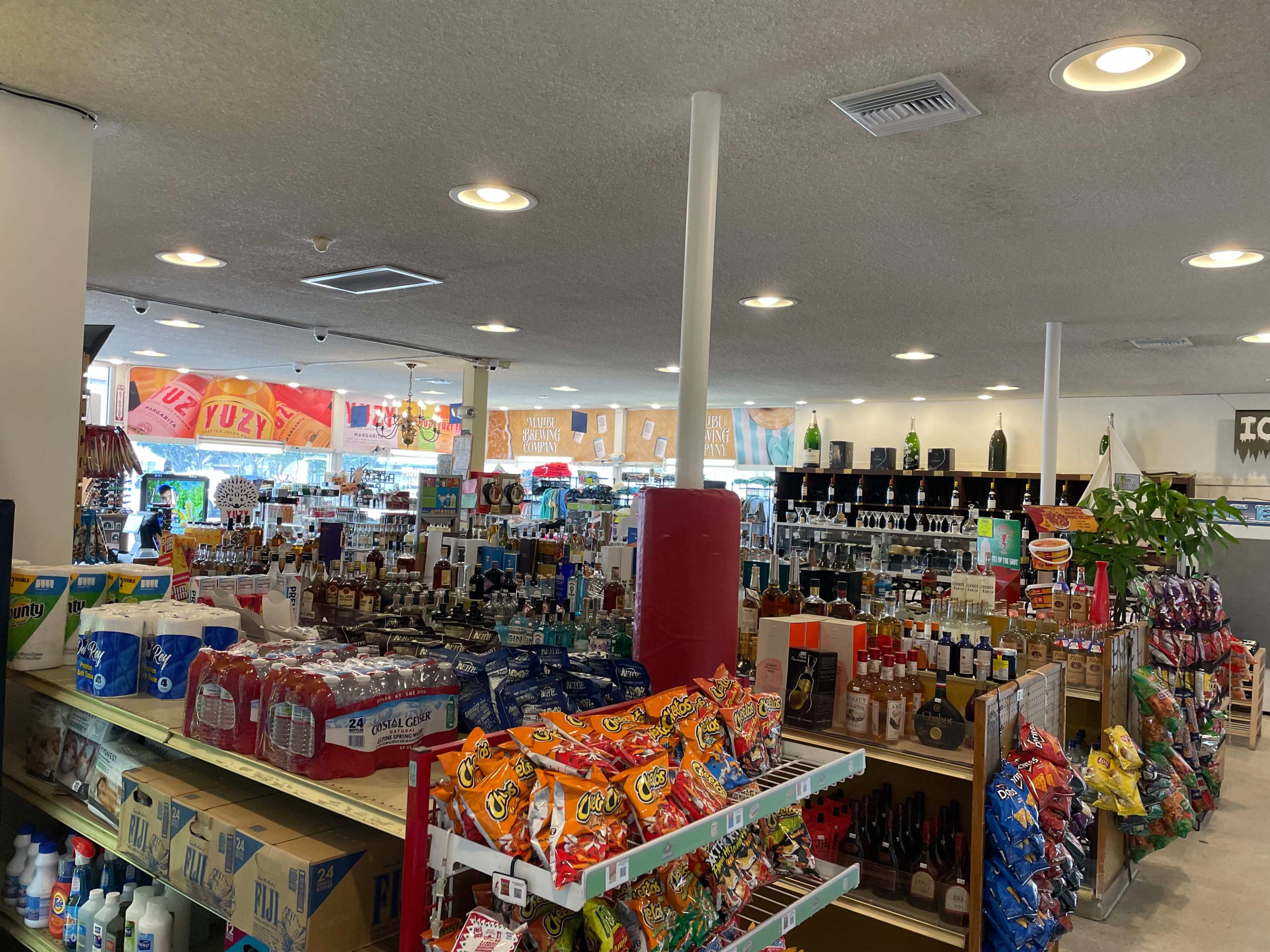The image shows the interior of a convenience store filled with shelves of snacks, beverages, and alcohol, with bright overhead lighting and promotional signage visible in the background.