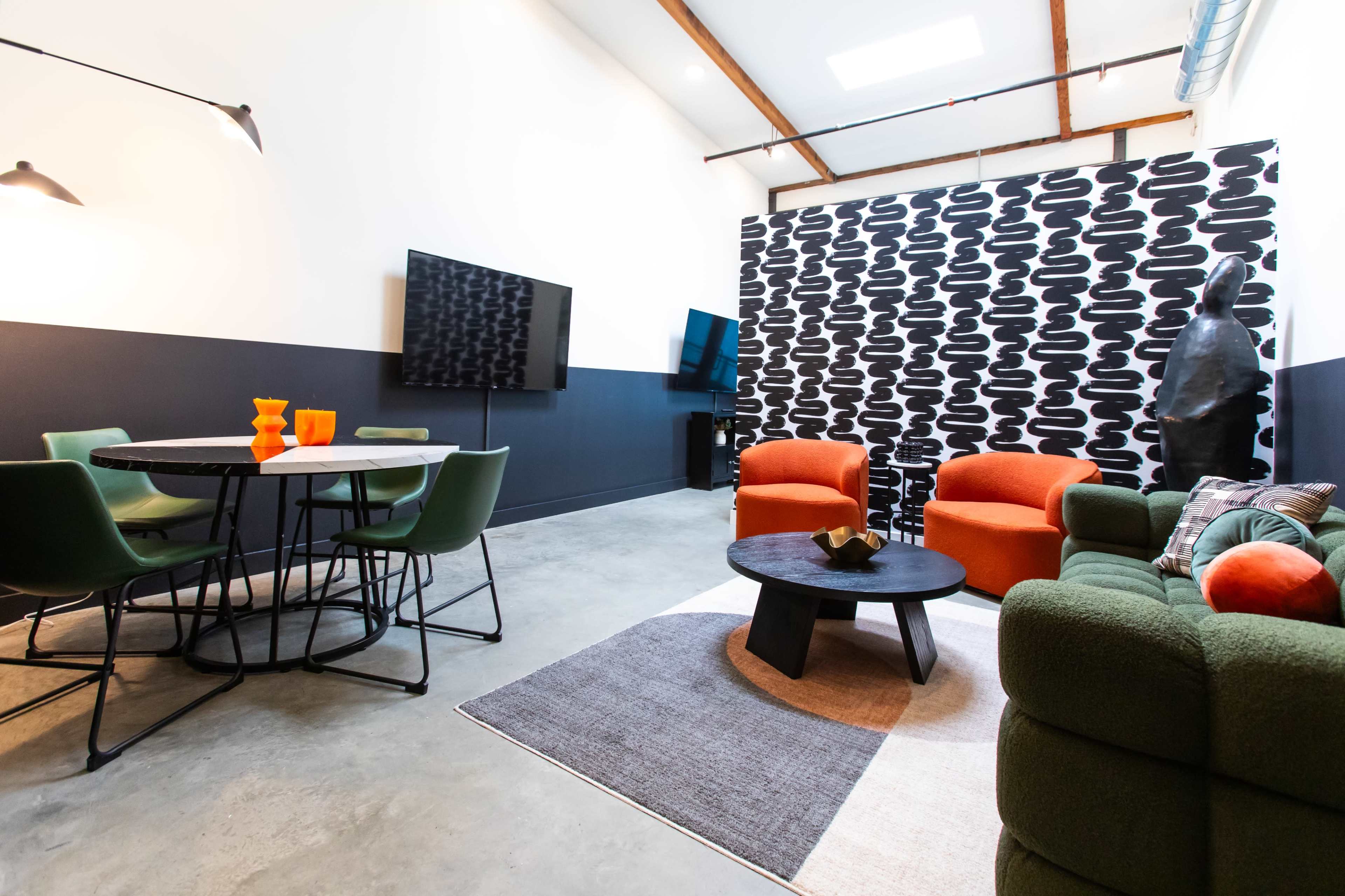 The image shows a modern living space featuring a round dining table with green chairs, a seating area with orange and green sofas, and a patterned accent wall.