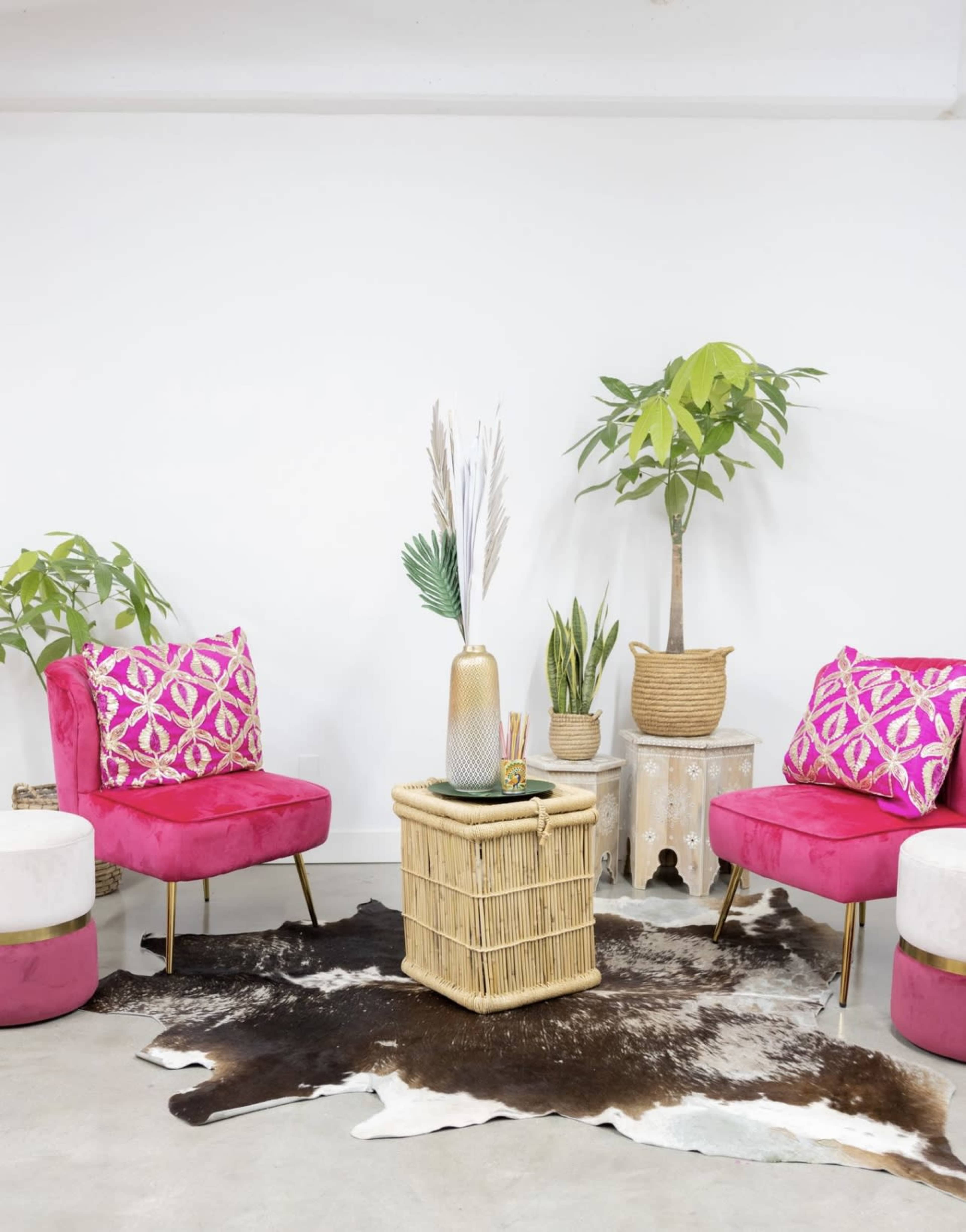 The image depicts two pink chairs with patterned cushions flanking a wicker coffee table, surrounded by plants and a cowhide rug in a bright, minimalist room.