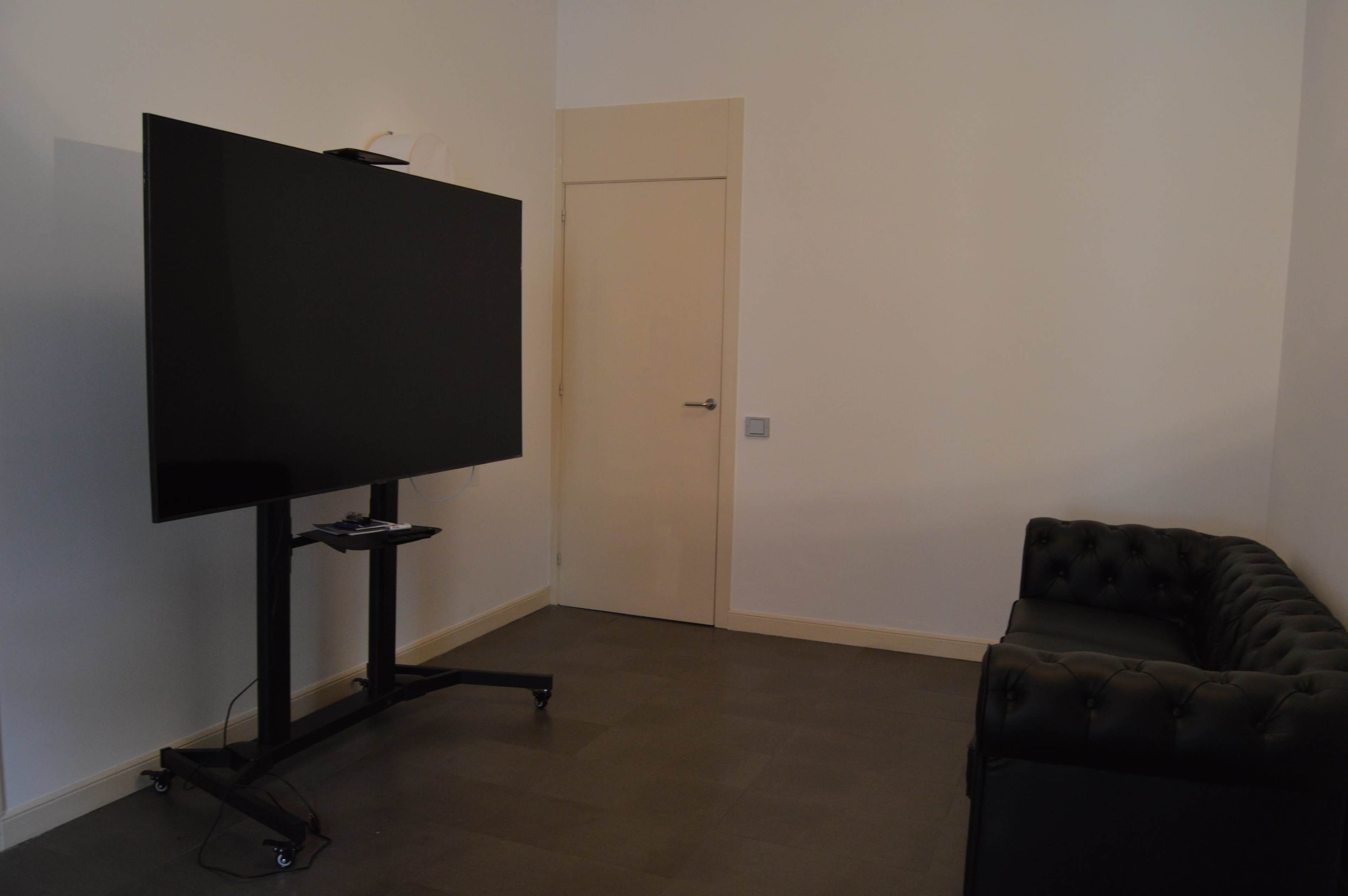 A minimalistic room with a large flat-screen TV mounted on a stand and a black leather couch facing it, alongside a closed door.