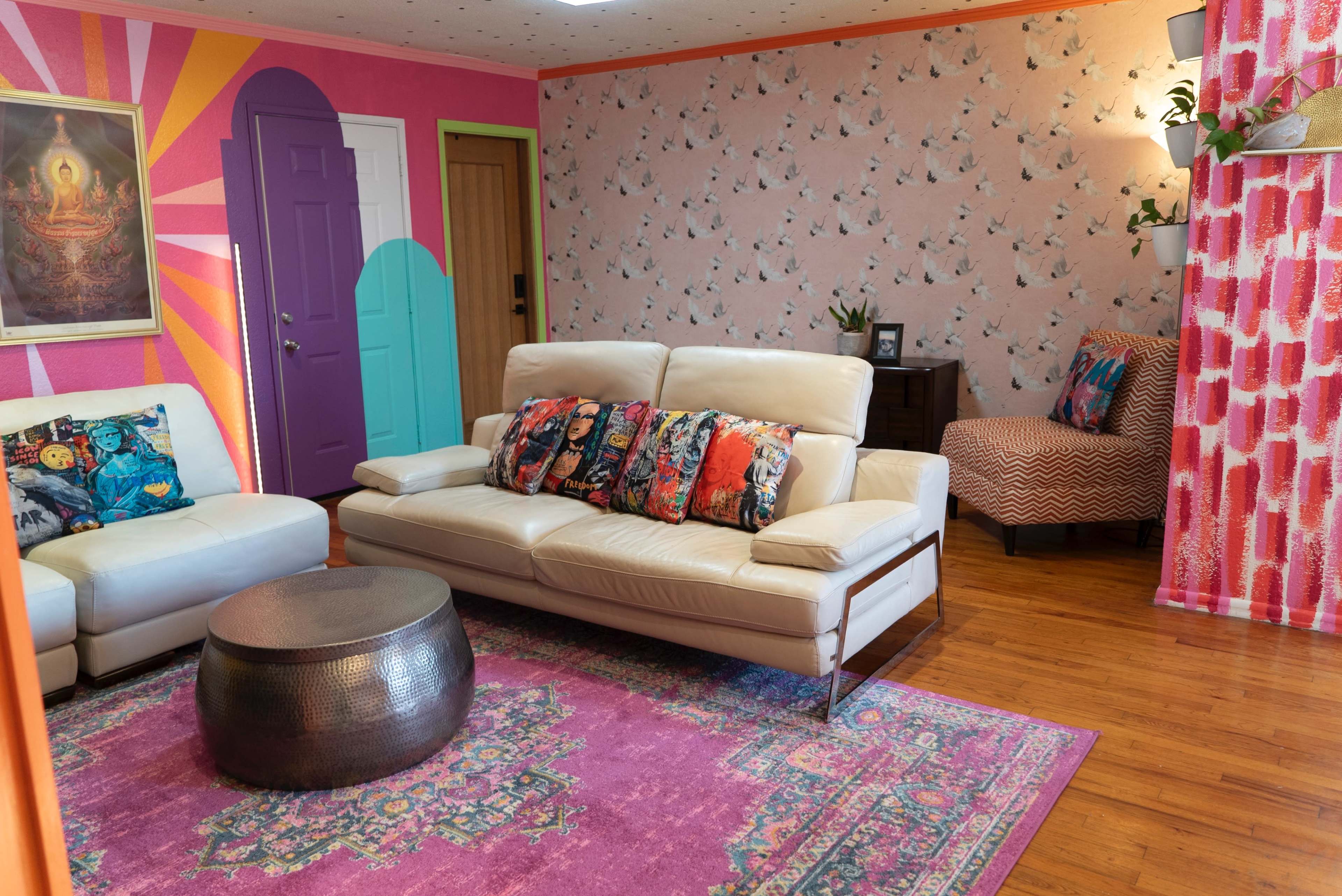 The image shows a brightly colored living room with a white leather sofa, a circular metal coffee table, and patterned walls featuring a mix of wallpaper designs and a colorful door.