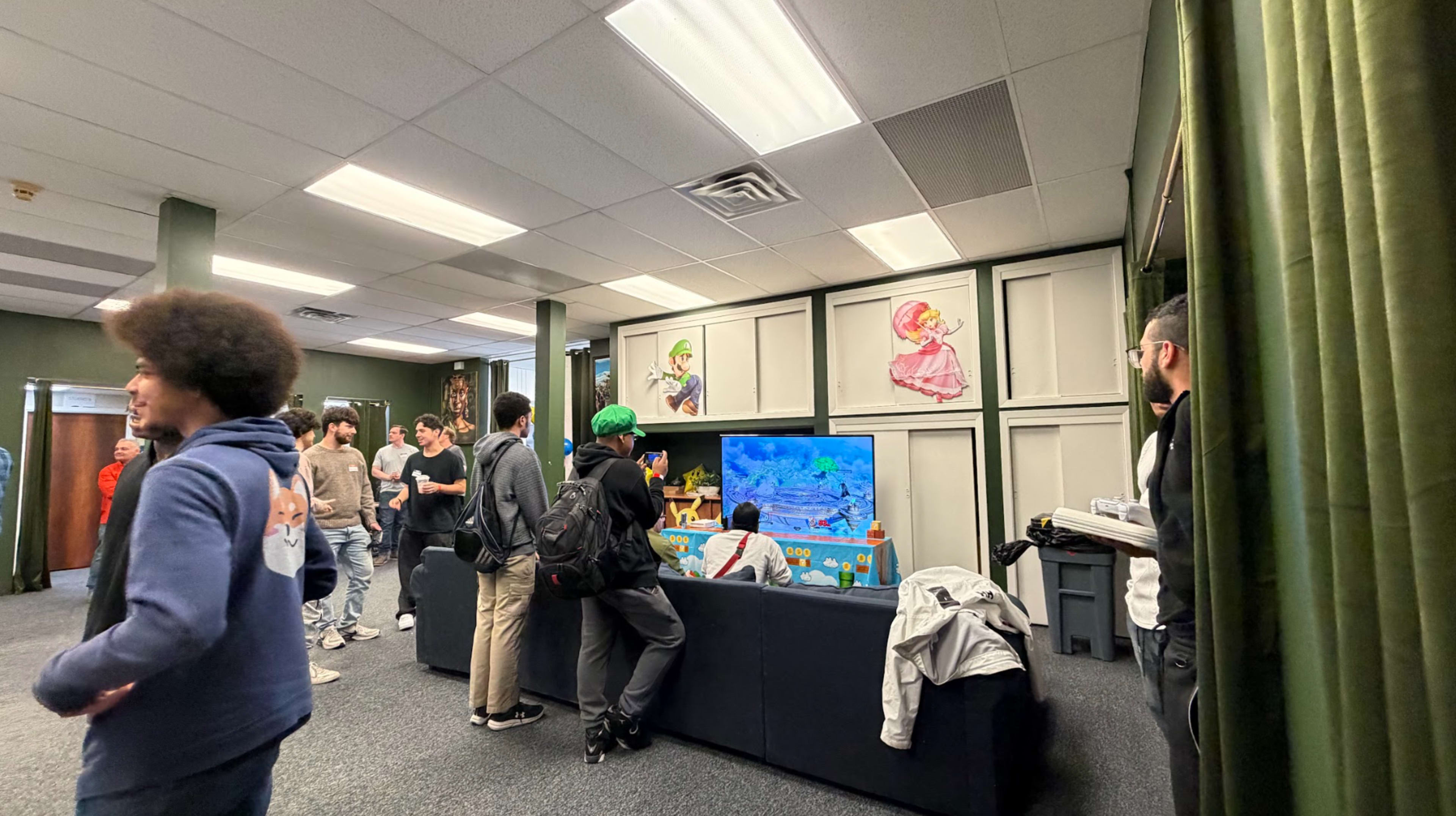 A group of people gathers in a room with a television displaying a video game, while others chat nearby.