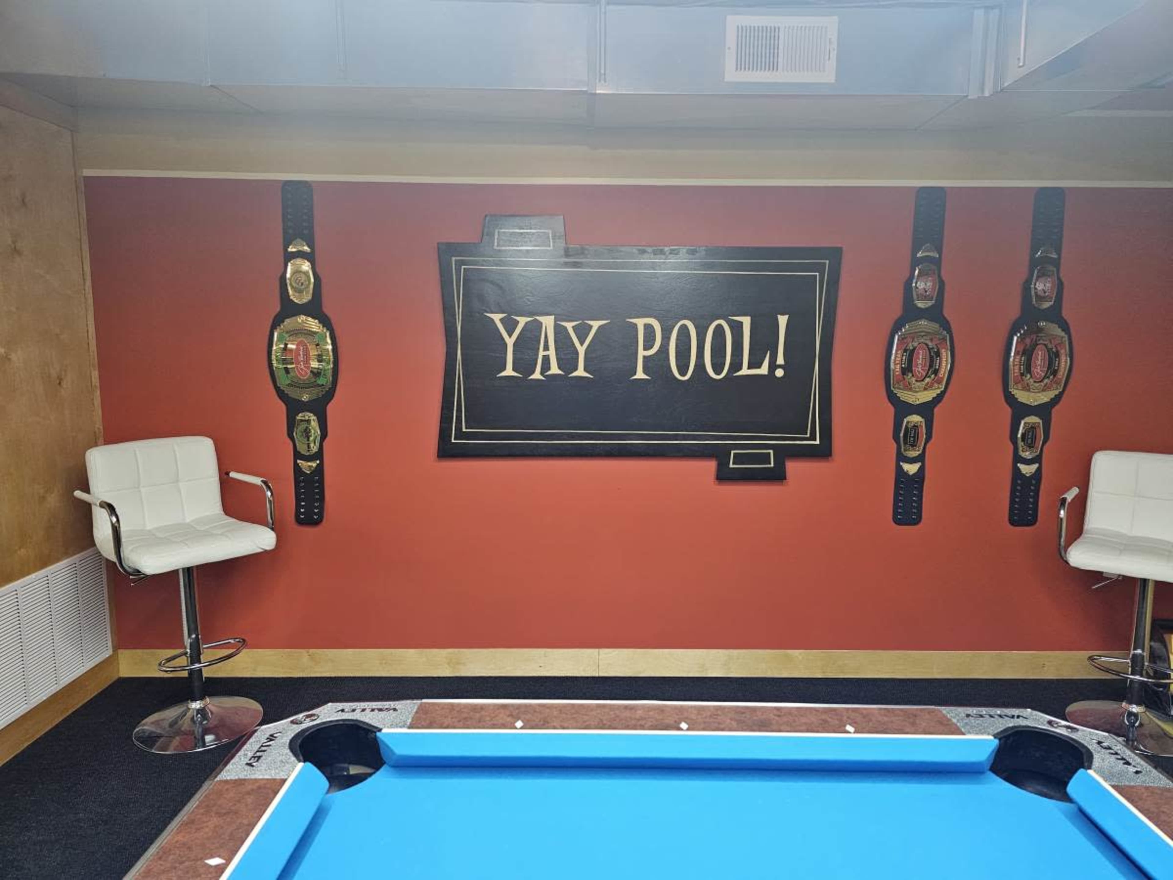 The image shows a vibrant room with a blue pool table, two white bar stools, and a wall decorated with the text "YAY POOL!" and three championship-style belt designs.