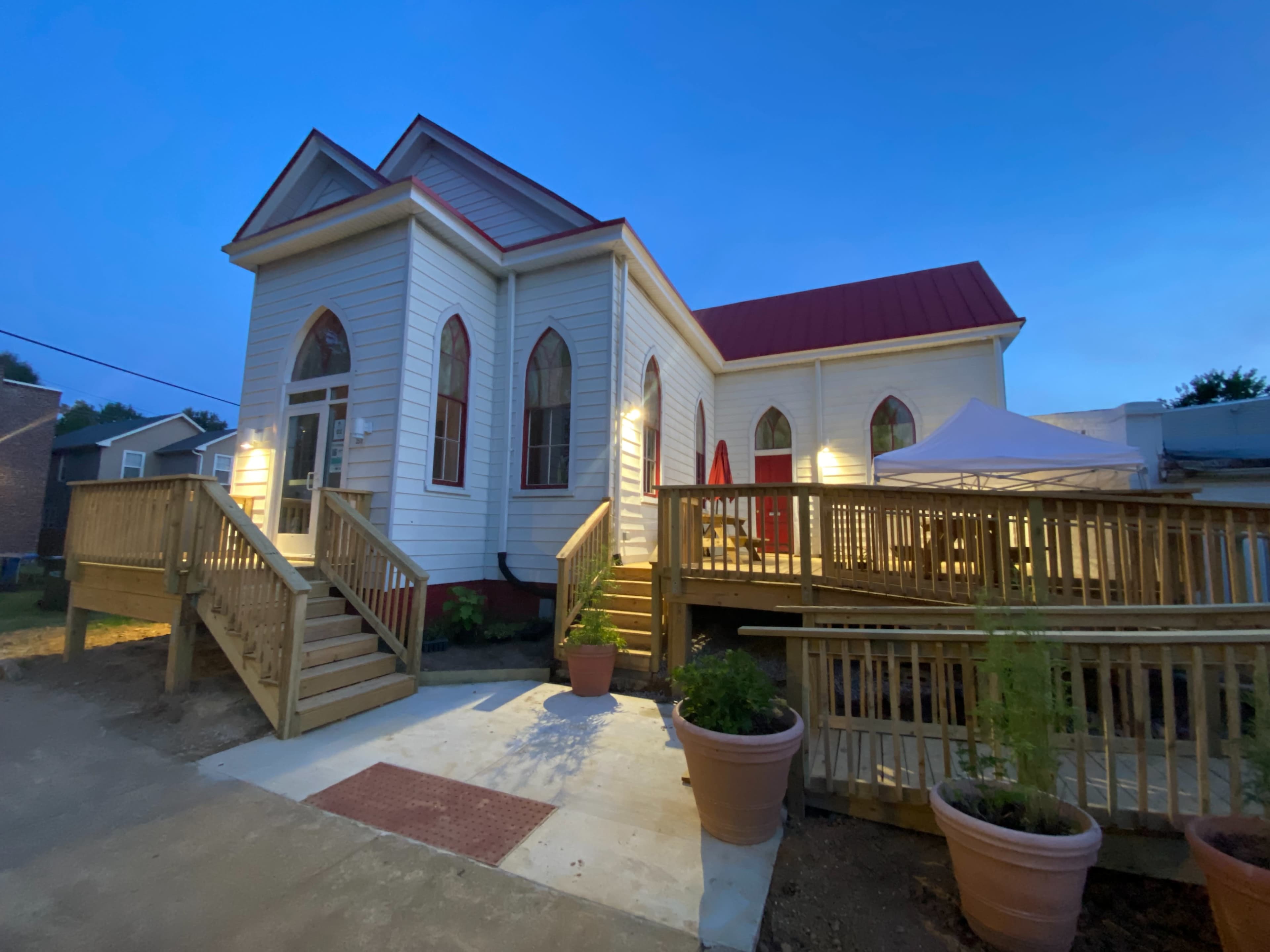 The image shows a house with a distinctive architectural style featuring pointed windows, a red metal roof, and a wooden ramp leading to a spacious deck.