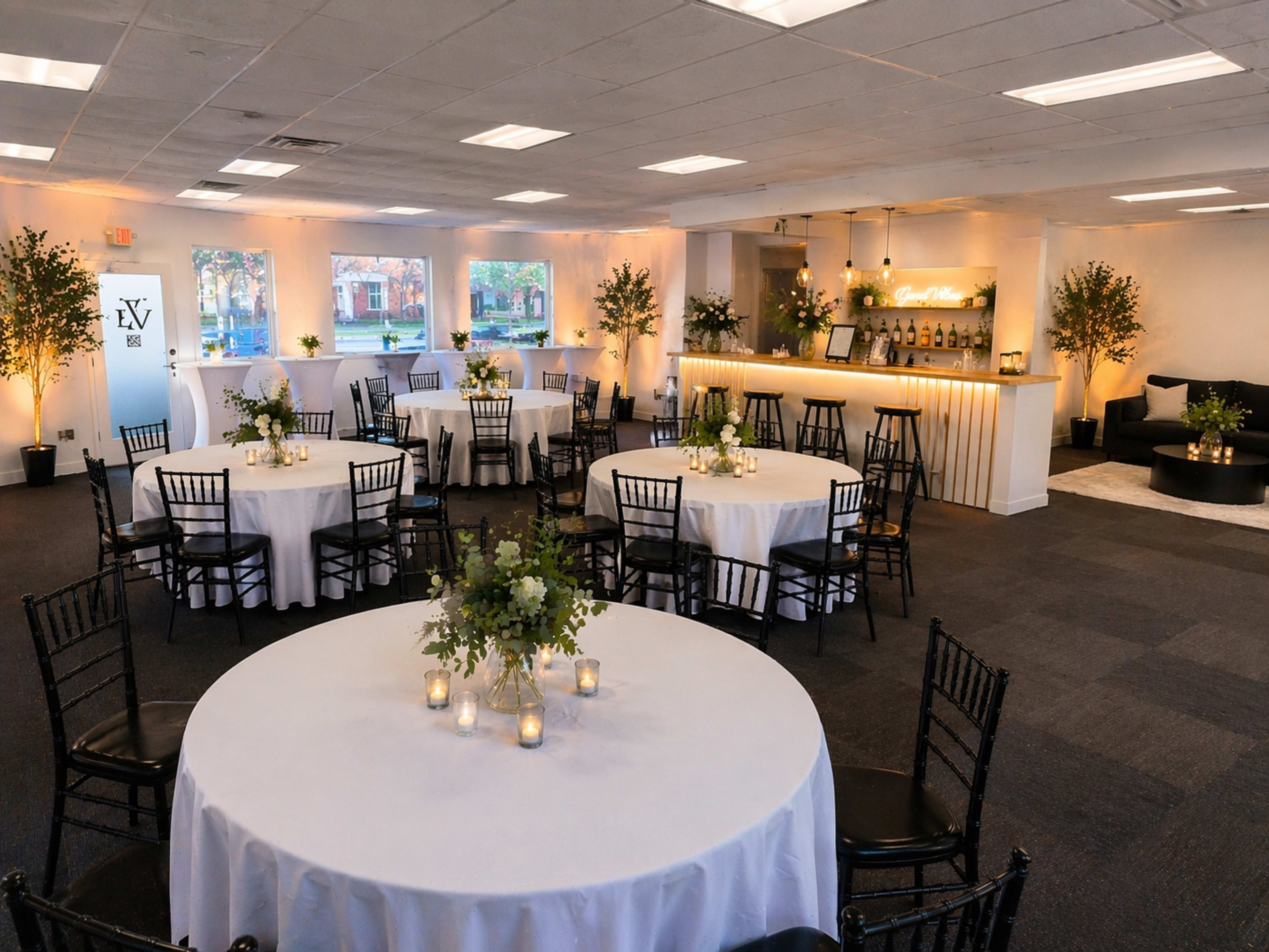 A spacious event venue features several round tables set with white tablecloths, centerpieces of greenery, and lit candles, with a bar area visible in the background.