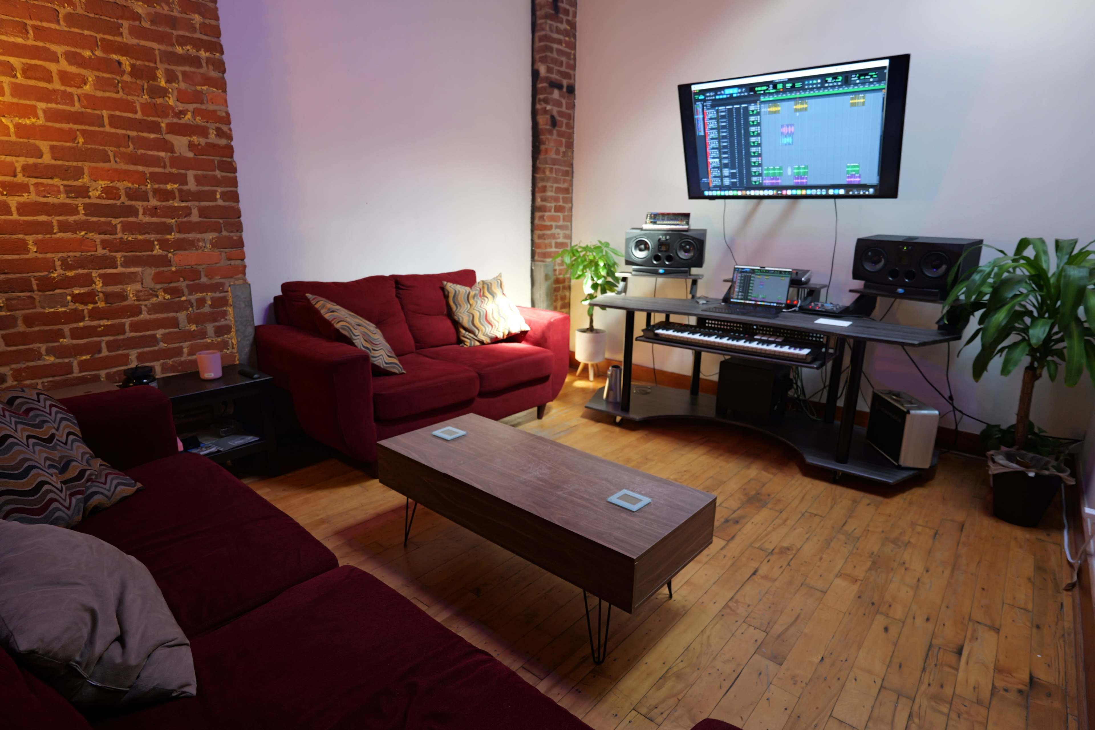 A cozy music studio features red couches, a wooden coffee table, and a workspace with a computer, speakers, and a keyboard against a brick wall.