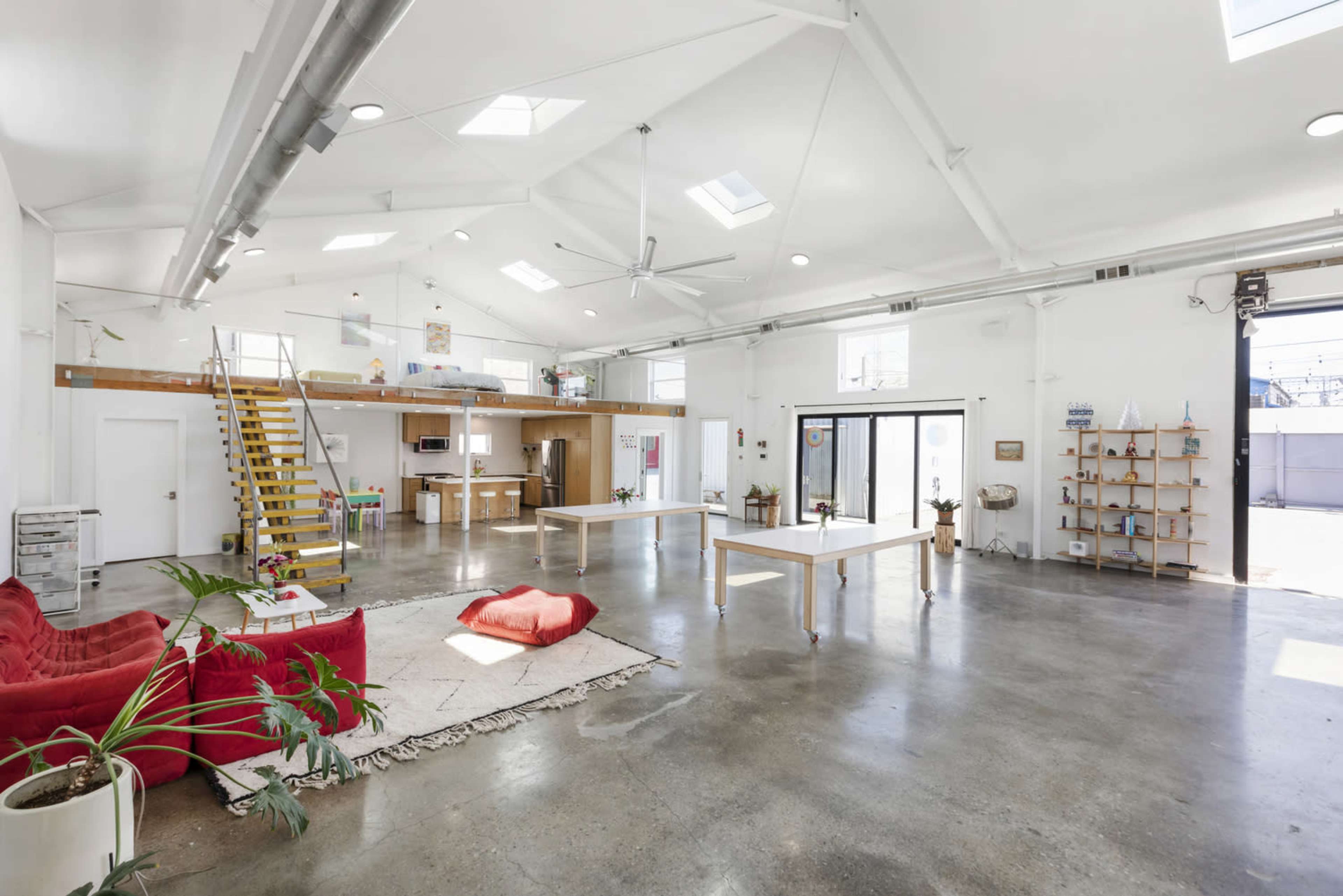 The image shows a spacious, well-lit interior of a modern studio with high ceilings, large windows, and minimalist furniture, including a red couch and white tables.