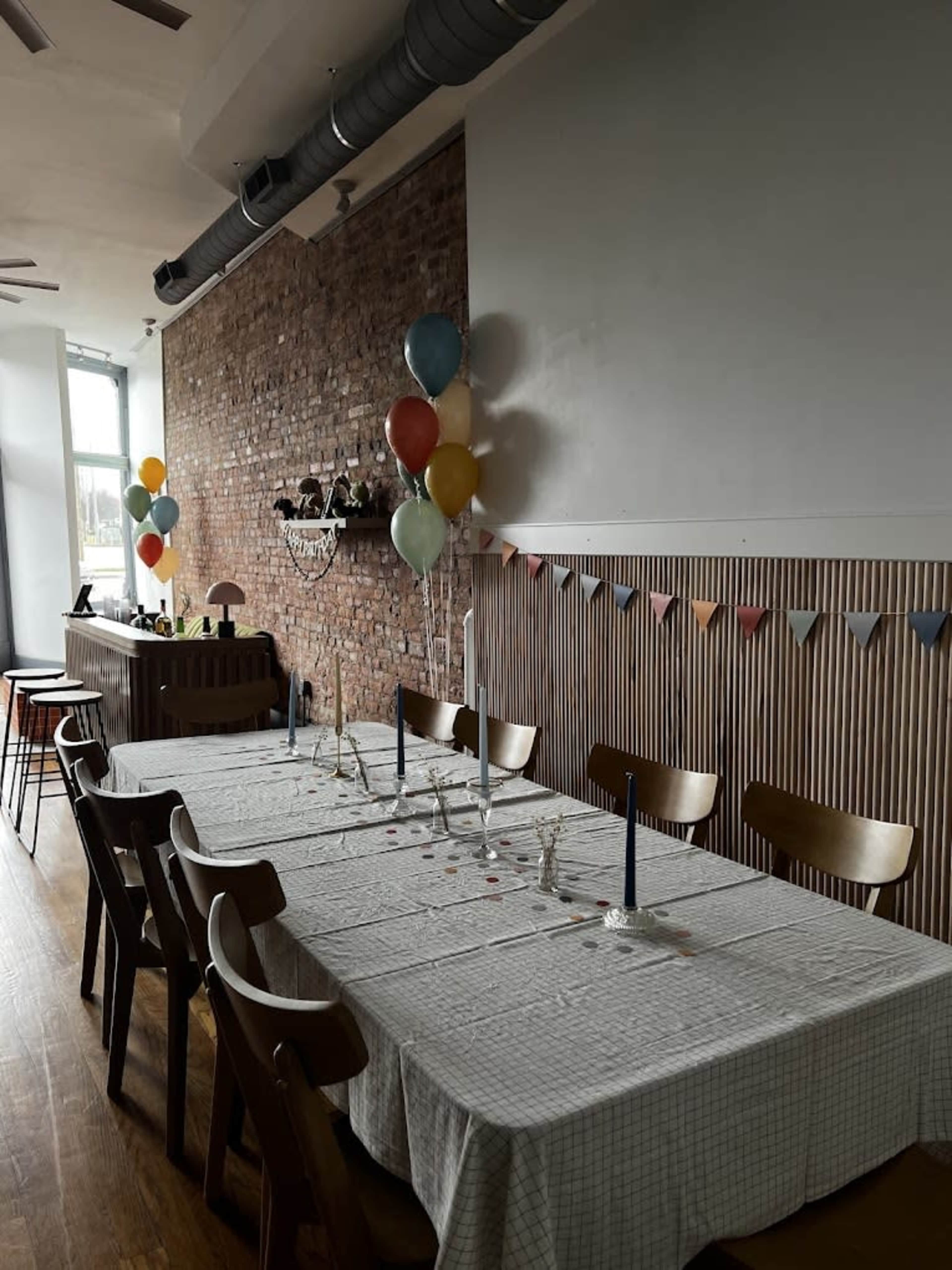 A long dining table set with candles and a checkered tablecloth is positioned against a brick wall, decorated with balloons and a bunting banner.