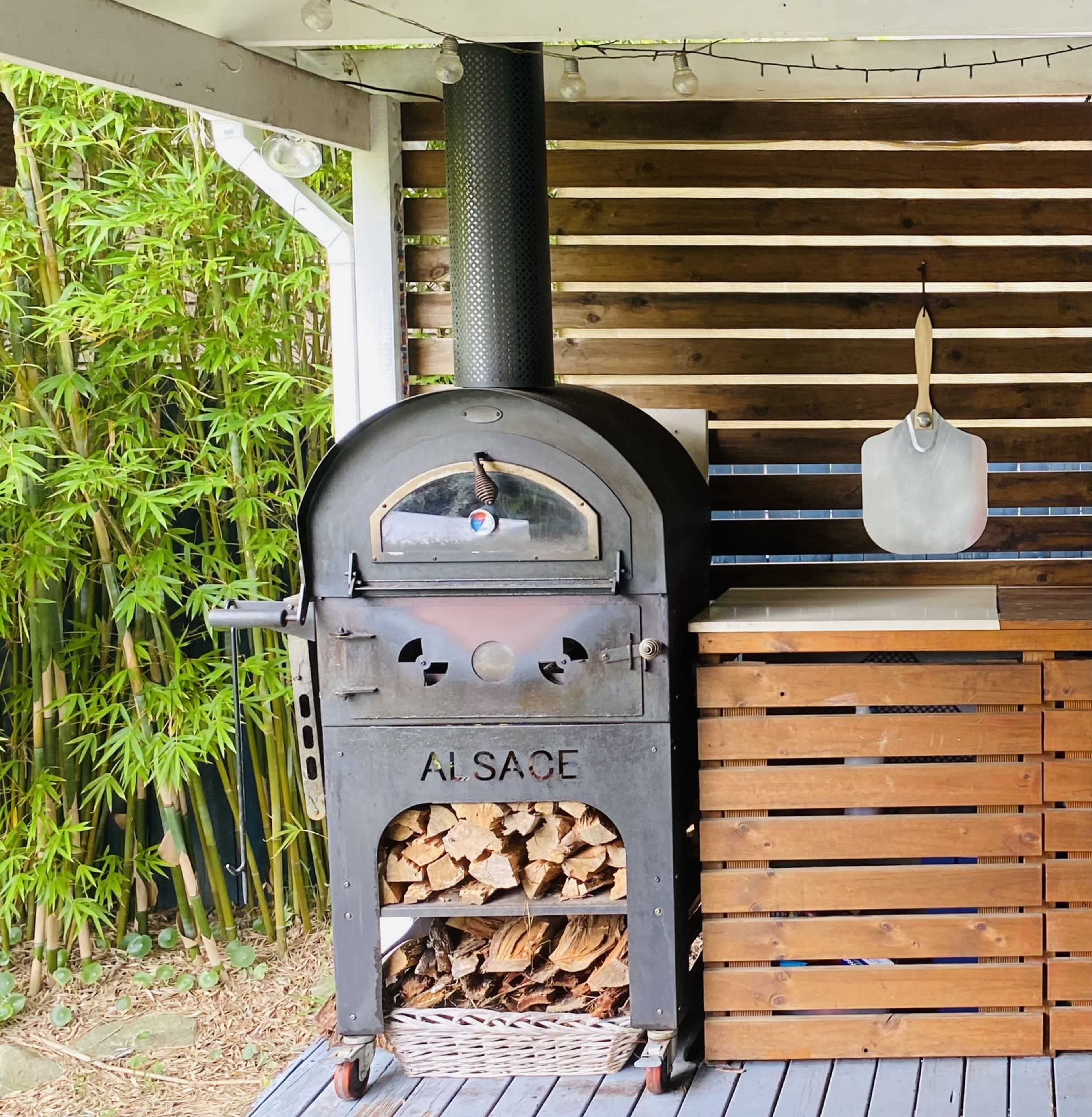 The image shows a black outdoor pizza oven with a wood storage area and a wooden counter, surrounded by bamboo plants.