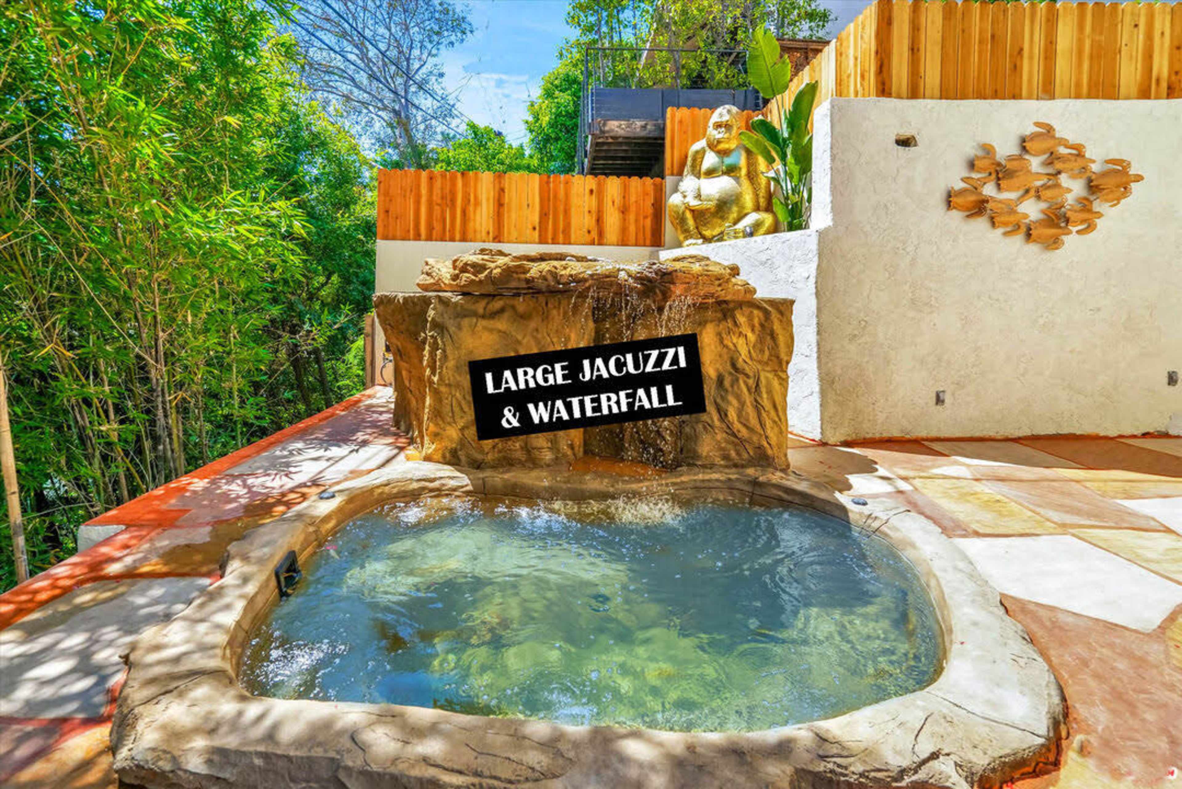 The image shows a large jacuzzi surrounded by rocks and a waterfall, with a statue seated nearby in a lush outdoor setting.