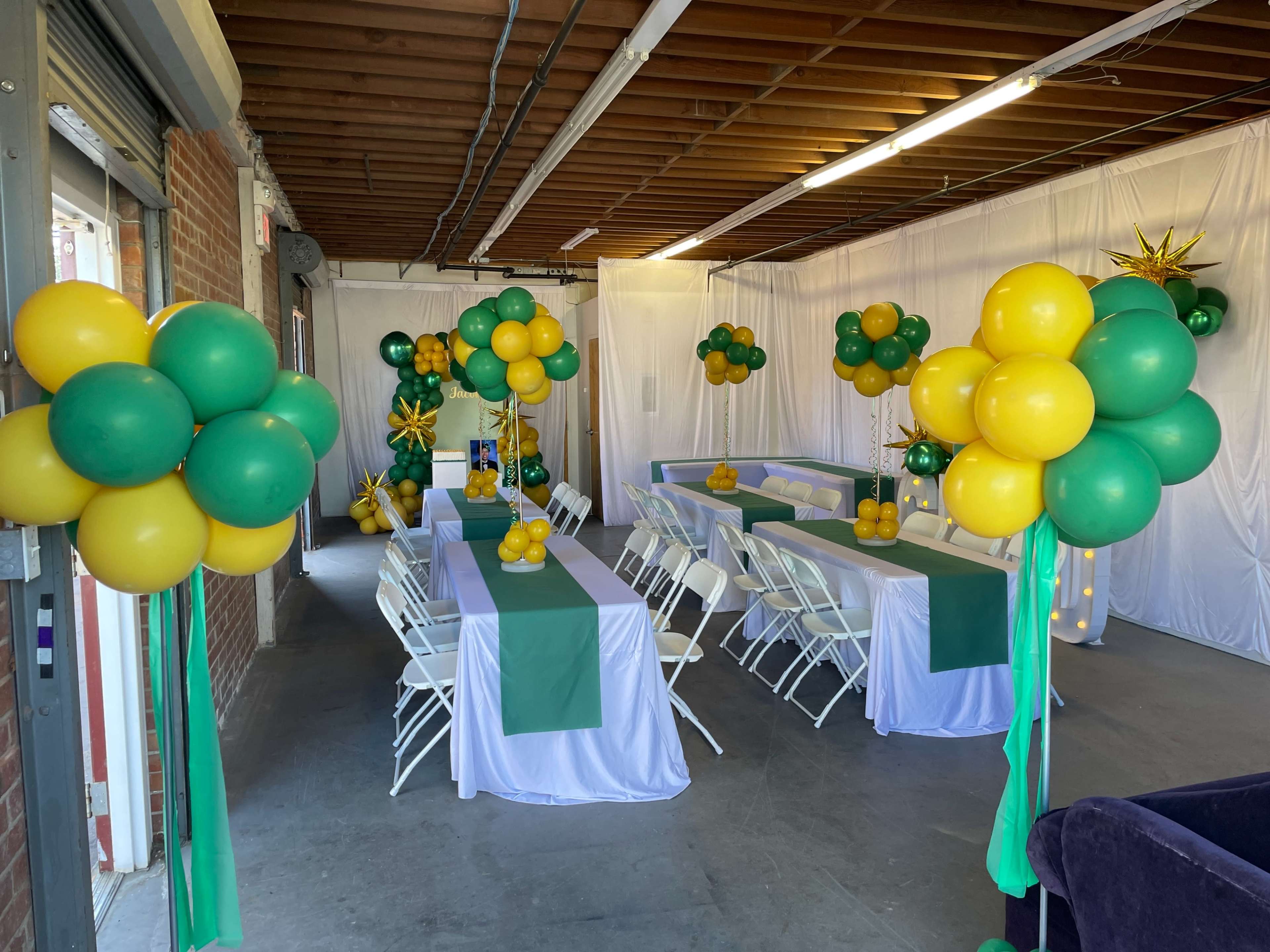 A decorated event space with tables covered in white and green tablecloths, surrounded by balloon arrangements in yellow and green.