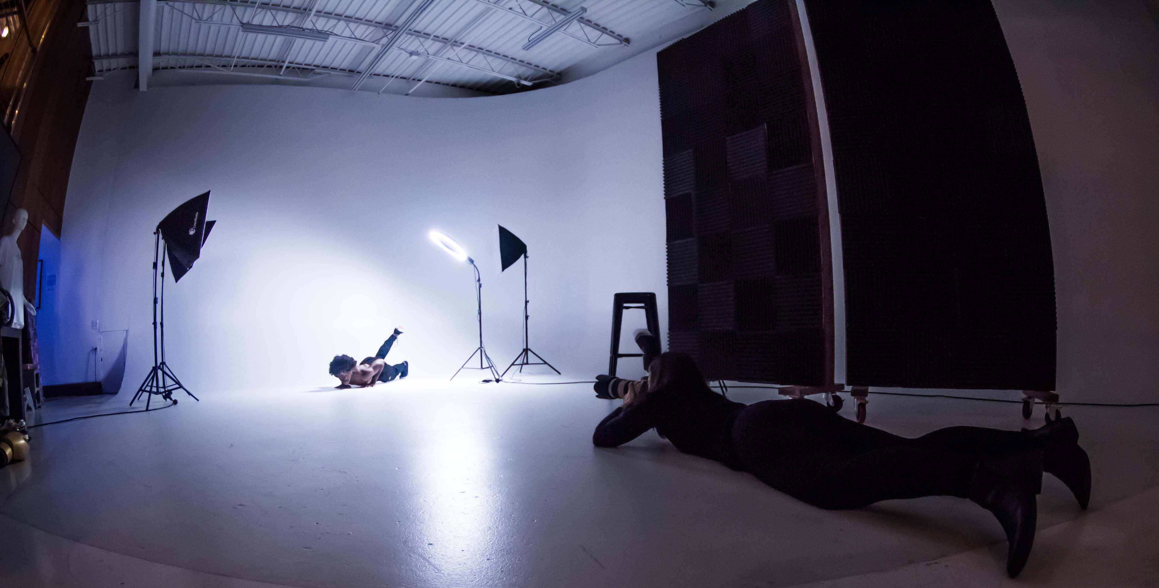 A photographer and a model are positioned on the floor of a studio, surrounded by lighting equipment and set against a white backdrop.