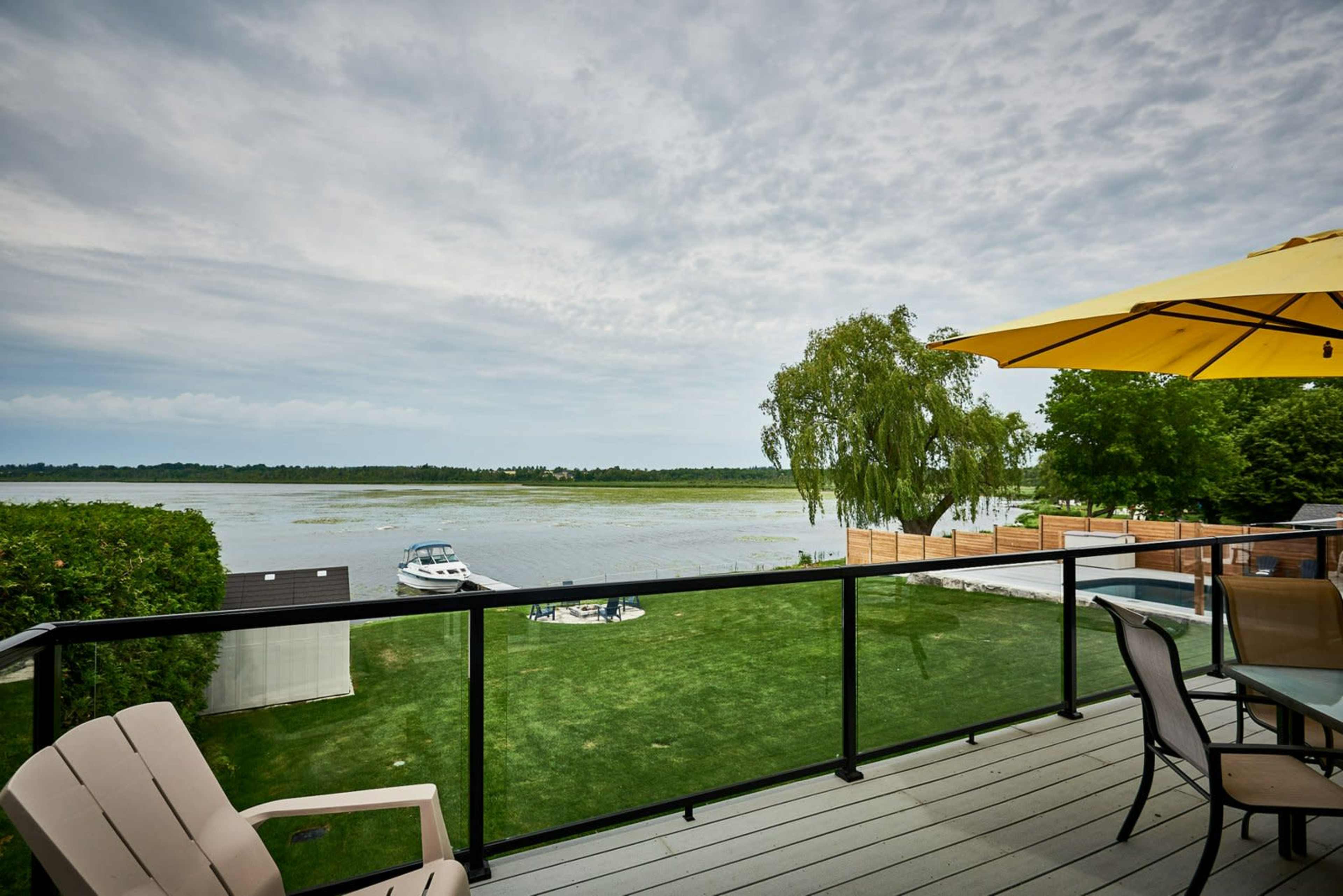 A lake view from a balcony featuring a yellow umbrella and several wooden chairs, with a boat docked nearby.