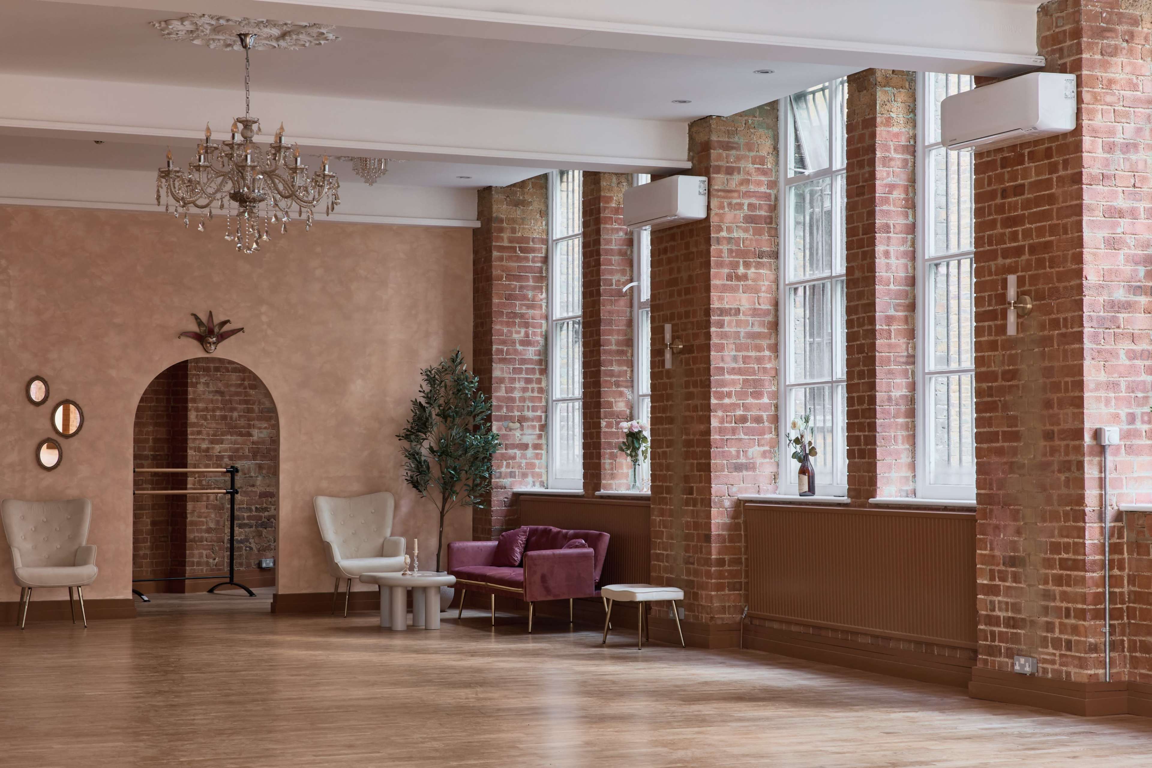 A spacious interior room features large windows, exposed brick walls, elegant light fixtures, and modern furniture arranged around a wooden floor.