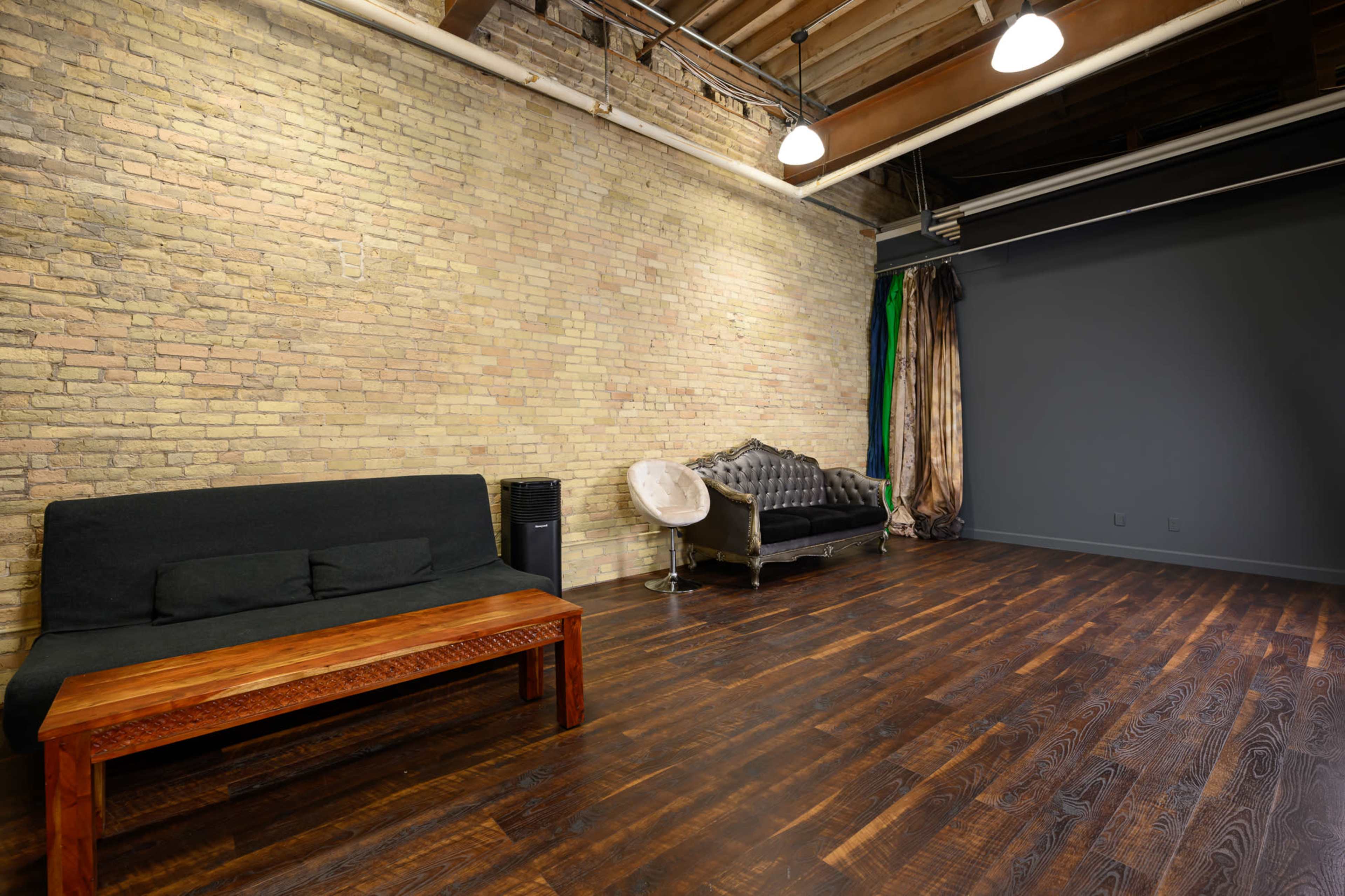 The image shows a spacious room with exposed brick walls, dark wooden flooring, a black sofa, and a silver upholstered couch.