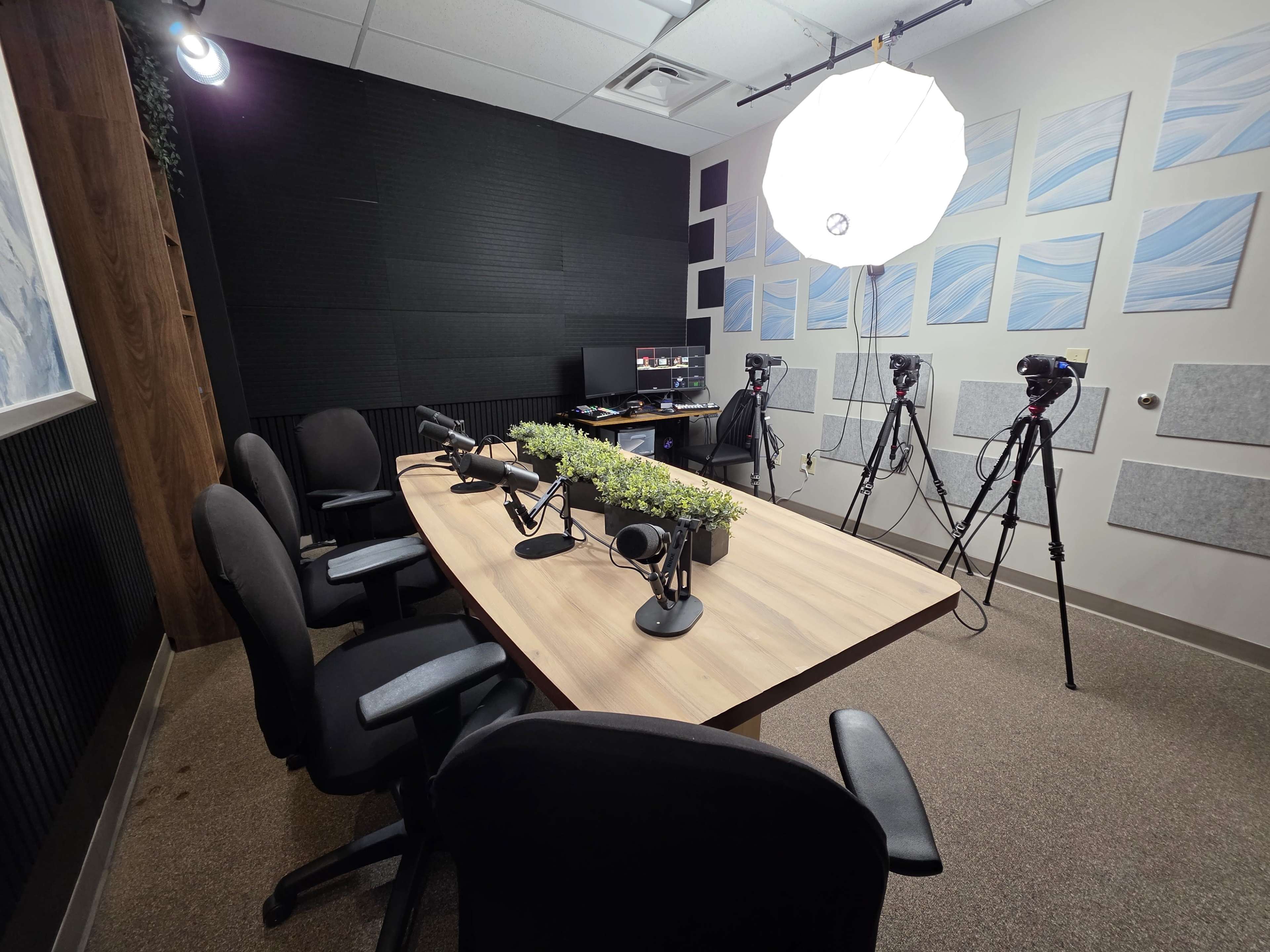 The image shows a podcast recording studio with a long table, four chairs, microphones, plants, and camera equipment positioned for video recording.